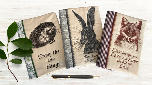 three embroidered notebook covers, one with a hedghog, one with a hare and one with a fox