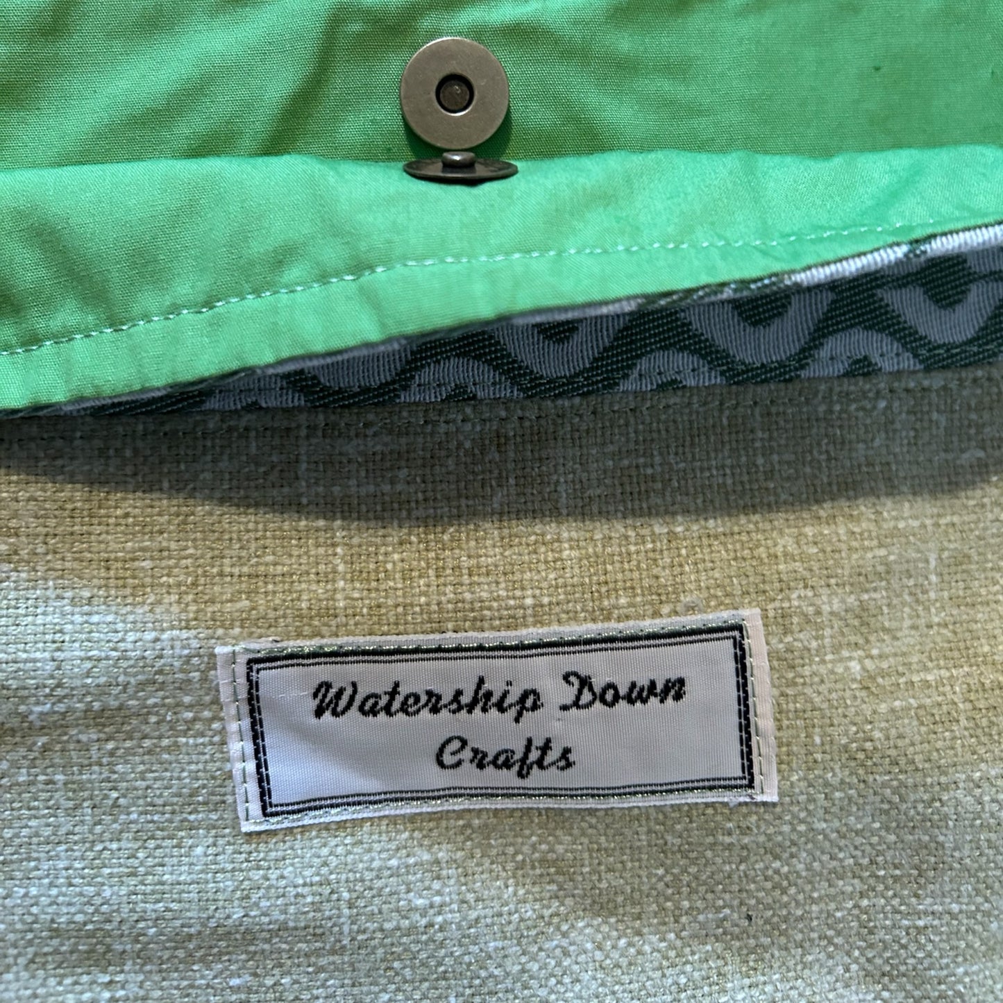 A close-up view of a handmade sage green bag's interior, highlighting a bright mint green lining and a secure magnetic snap closure. The lining edge features a subtle geometric cream and green pattern for added detail. A "Watership Down Crafts" label is stitched onto the sage green fabric, showcasing the handcrafted branding. The image emphasizes the bag's thoughtful design and functional features.