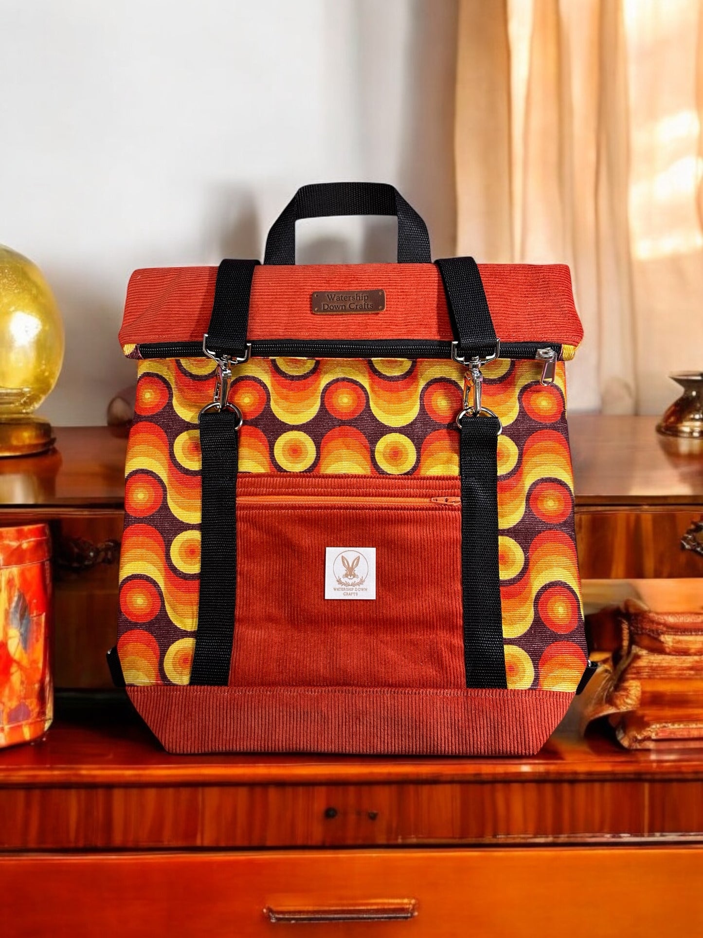 A vibrant orange roll-top backpack with a retro-inspired design displayed on a wooden dresser. The fabric has a bold pattern with concentric circles in shades of orange, yellow, and red. The backpack features black straps, silver hardware, and a front pocket adorned with the "Watership Down Crafts" logo on a white tag. A small, leather brand label is positioned on the top flap. The warm, vintage setting includes golden decorative items that complement the retro style of the bag.