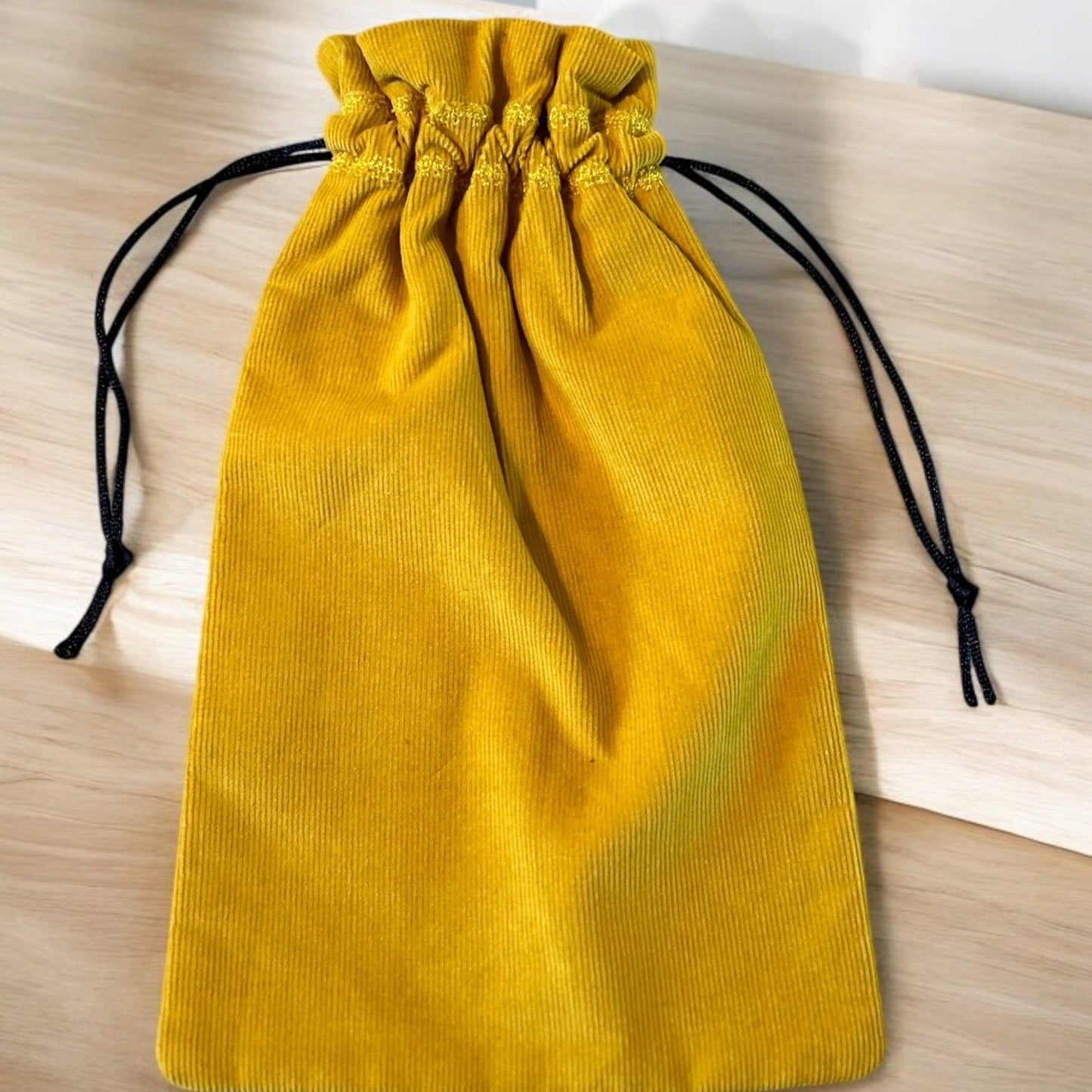 A handmade drawstring bag crafted from mustard yellow corduroy fabric, showcasing a soft textured appearance. The bag is cinched with black drawstring cords for a contrasting touch, combining functionality with bold color. It is displayed on a light wooden surface, highlighting the richness of the fabric.