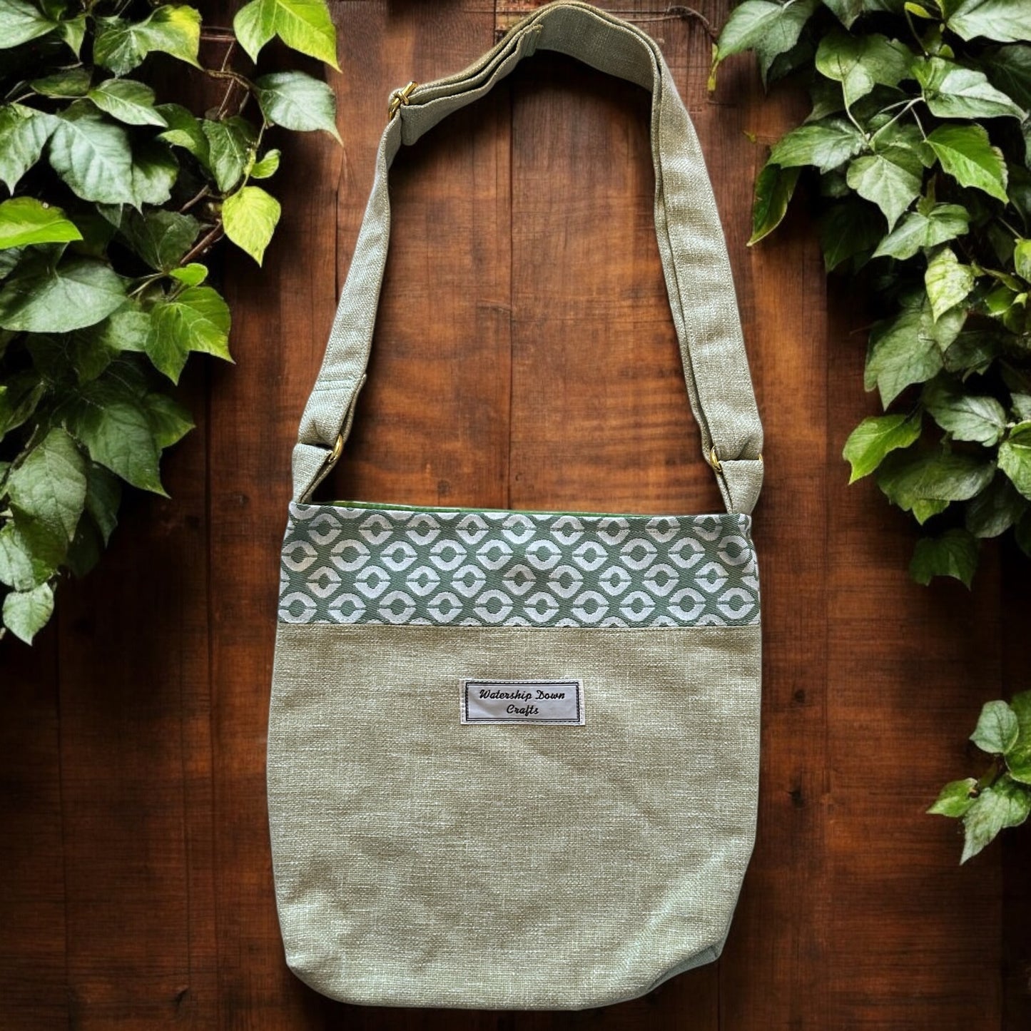 A handmade crossbody bag with a soft sage green base fabric and a decorative geometric cream and green patterned band across the top. The bag features an adjustable strap in matching sage green fabric with gold-tone hardware. A "Watership Down Crafts" label is prominently stitched on the front, adding a personalized touch. It is displayed against a warm wooden backdrop surrounded by green ivy, creating a natural and rustic aesthetic.