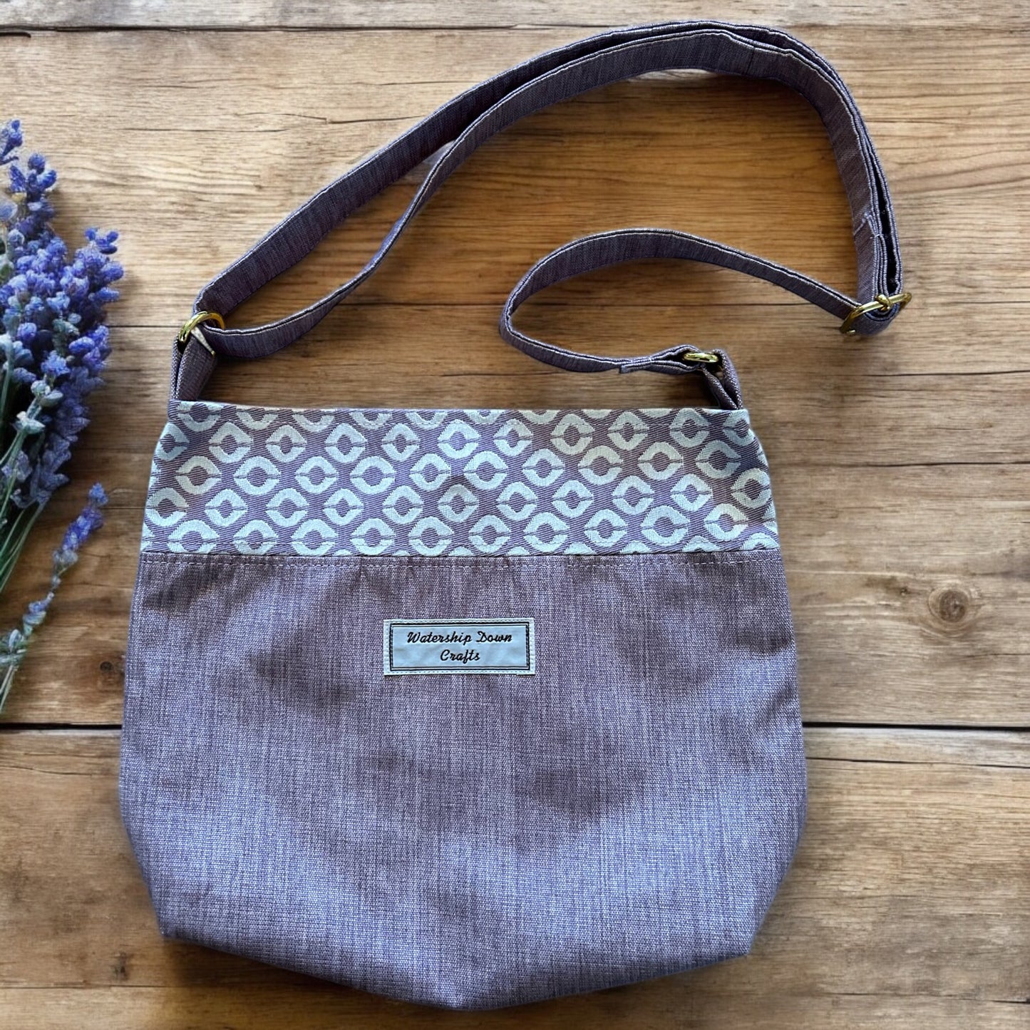 A handmade crossbody bag crafted with a soft lilac fabric base and a patterned band of geometric cream and lilac accents across the top. The bag features an adjustable strap in matching fabric with gold-tone hardware. A "Watership Down Crafts" label is prominently sewn on the front, adding a personalized touch. The bag is displayed on a rustic wooden surface, with a sprig of lavender placed beside it, enhancing its delicate and sophisticated appeal.
