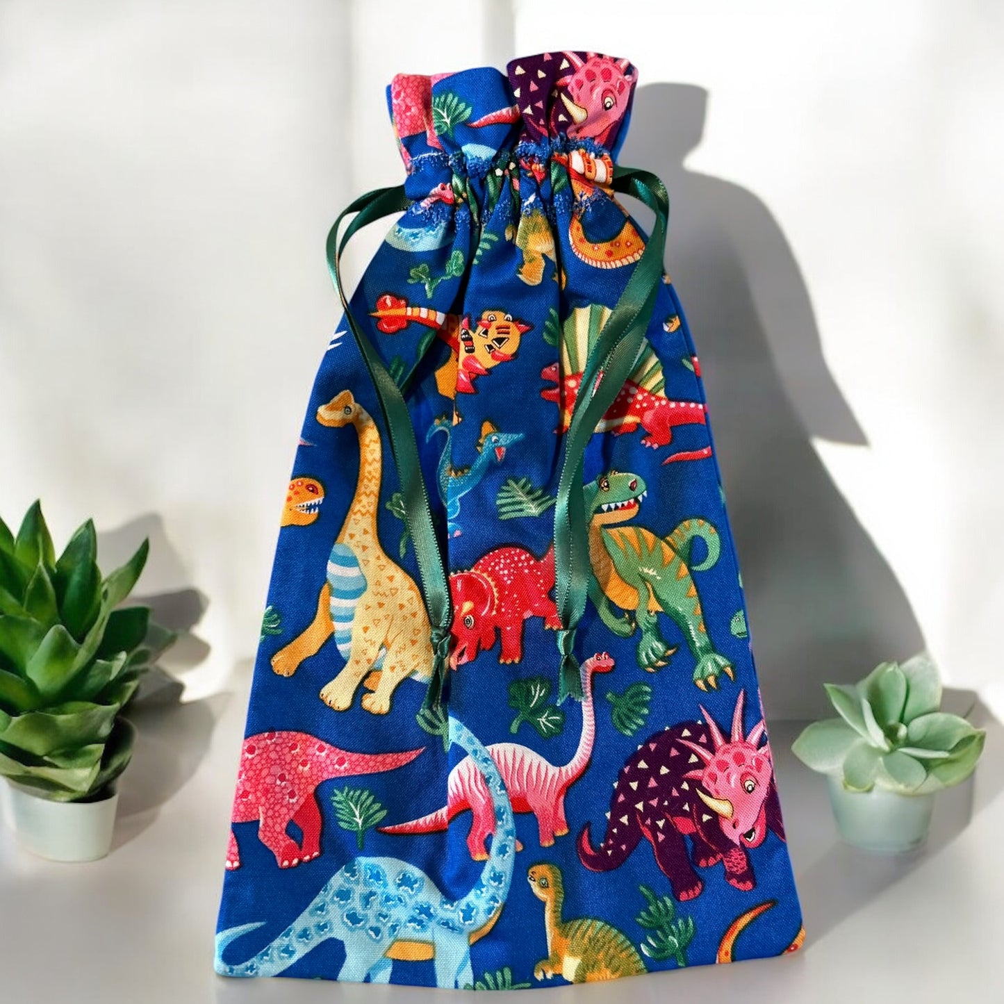 A vibrant handmade drawstring bag featuring a playful dinosaur print on a bright blue background. The colorful dinosaurs are detailed with cheerful expressions and various patterns, making the bag ideal for kids or dinosaur enthusiasts. It is cinched with green satin ribbon drawstrings and displayed with small succulent plants, adding a touch of greenery to the scene.