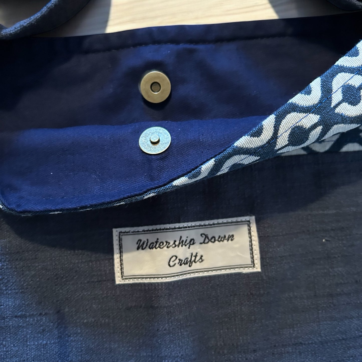 A close-up view of a handmade bag's interior, showcasing a magnetic snap closure for secure fastening. The lining features a solid navy blue fabric, while the edge highlights a geometric cream and navy pattern. A "Watership Down Crafts" label is prominently stitched on the inside, emphasizing the handcrafted detail and branding. The image captures the bag's craftsmanship and functional design elements.