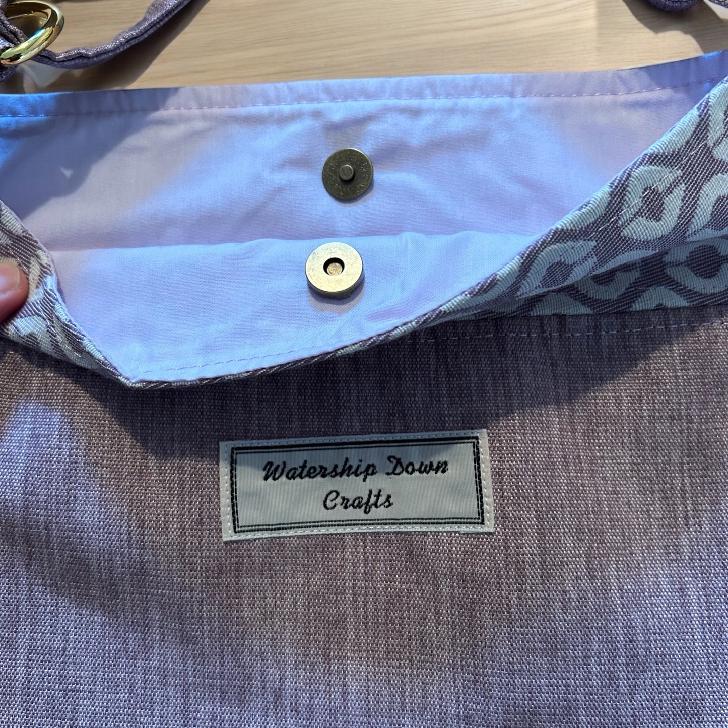 A close-up of the interior of a handmade lilac bag, showcasing a light lavender lining and a secure magnetic snap closure. The edge of the lining features a subtle geometric cream and lilac pattern, adding a decorative touch. A "Watership Down Crafts" label is stitched onto the lilac fabric, emphasizing the handcrafted and branded detail of the bag. The image highlights the craftsmanship and functionality of the design.
