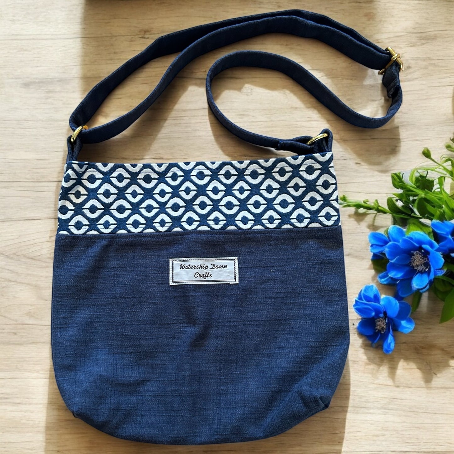 A stylish handmade crossbody bag featuring a navy blue base with a patterned band of geometric cream and navy accents across the top. The adjustable strap is made from matching navy fabric with gold-tone hardware. A "Watership Down Crafts" label is sewn on the front, adding a personal touch. The bag is displayed on a light wooden surface, complemented by vibrant blue flowers, creating a sophisticated yet fresh presentation.