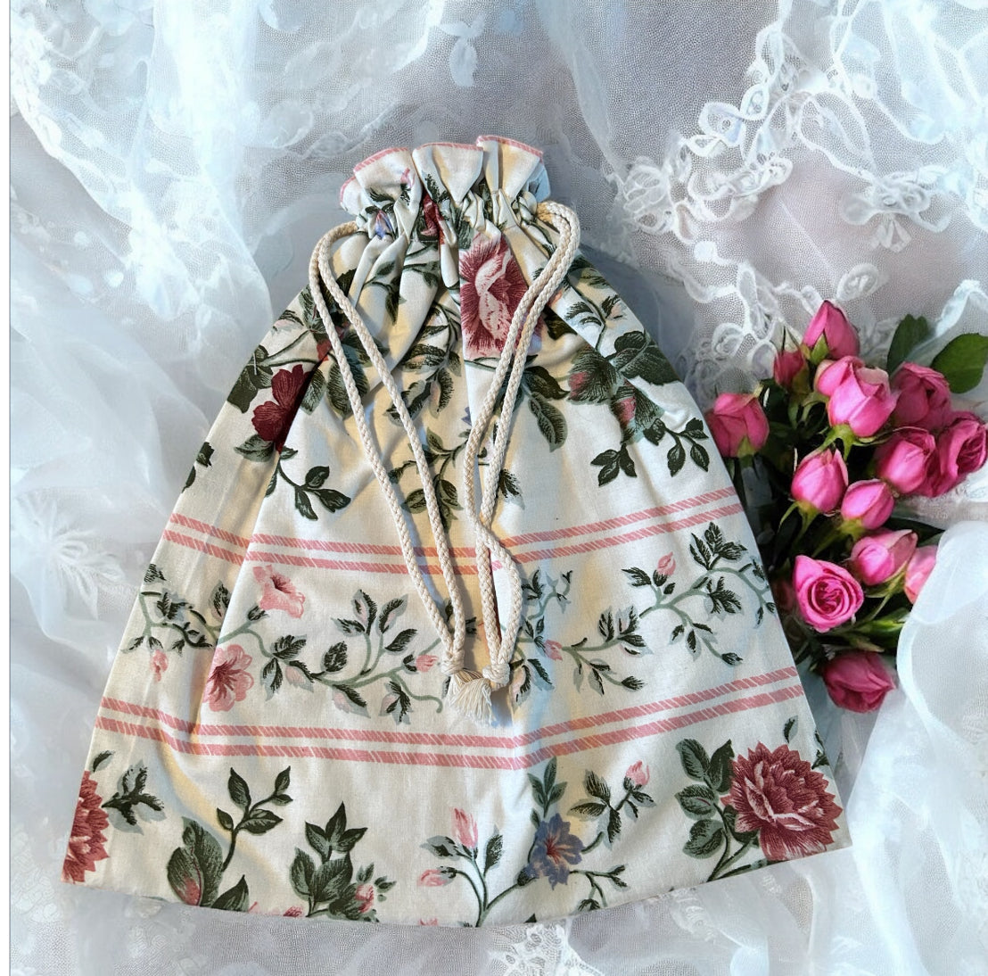 A handmade drawstring bag with a romantic floral design featuring pink and red roses with green leaves on a soft cream background, accented by delicate pink stripes. The bag is cinched with white rope drawstrings and displayed elegantly on a lace fabric, complemented by a bouquet of fresh pink roses. The overall aesthetic exudes charm and femininity.