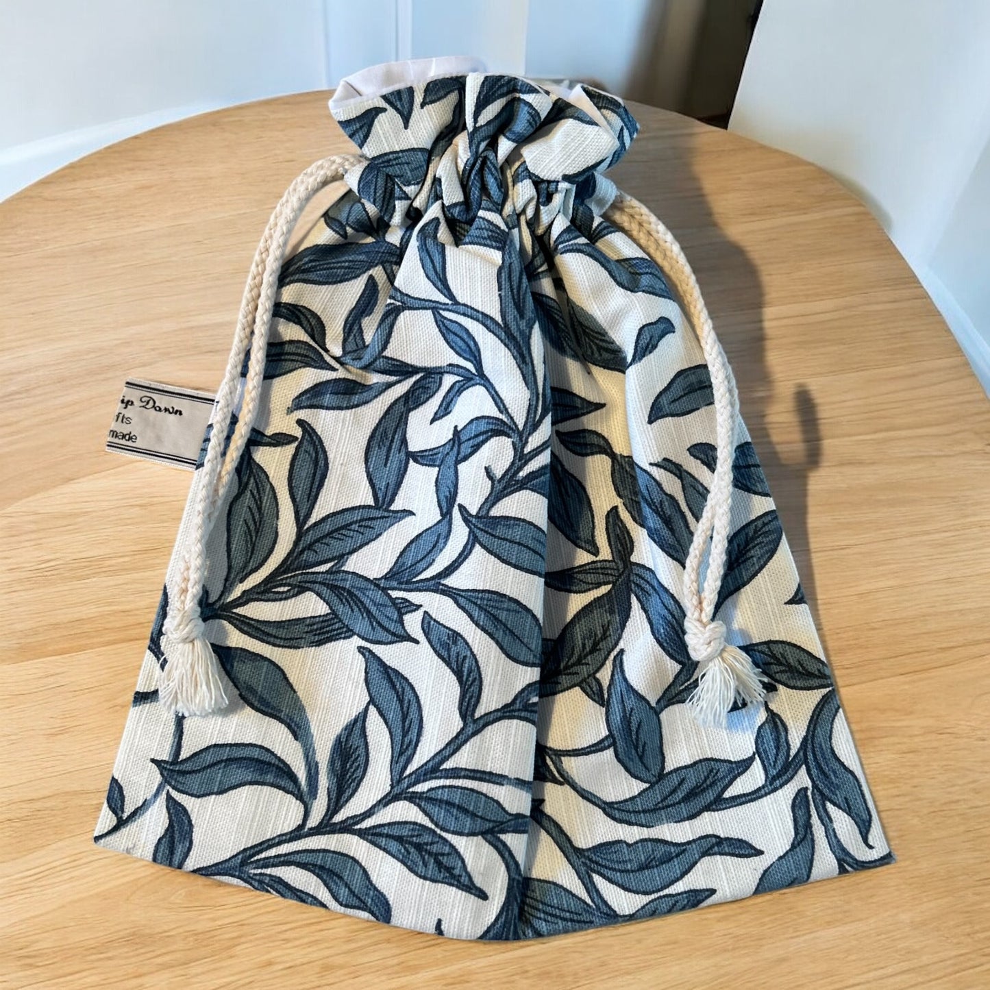 A handmade drawstring bag with a natural white base and an elegant blue leaf pattern. The bag features sturdy white rope drawstrings with knotted ends and tassels for added charm. It is displayed on a light wooden table, highlighting its simple yet stylish design. A small label from Watership Down Crafts is partially visible on the side.