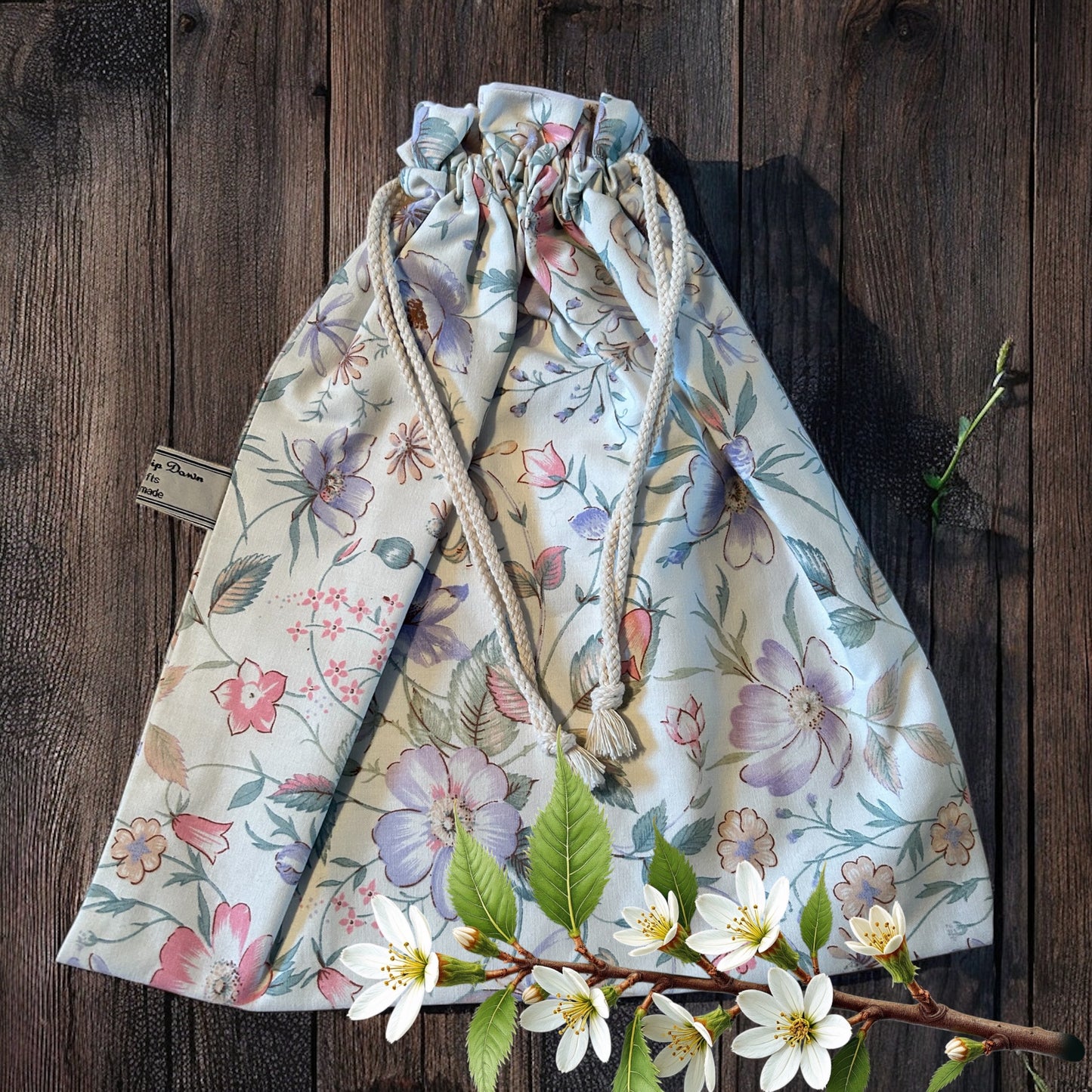 A charming handmade drawstring bag adorned with a delicate pastel floral pattern on a soft cream background. The bag features white rope drawstrings with knotted ends and is displayed on a rustic wooden surface. A sprig of blooming white flowers and green leaves adds a natural, fresh touch to the composition, highlighting the bag's spring-inspired design. A small "Watership Down Crafts" label is visible on the side.