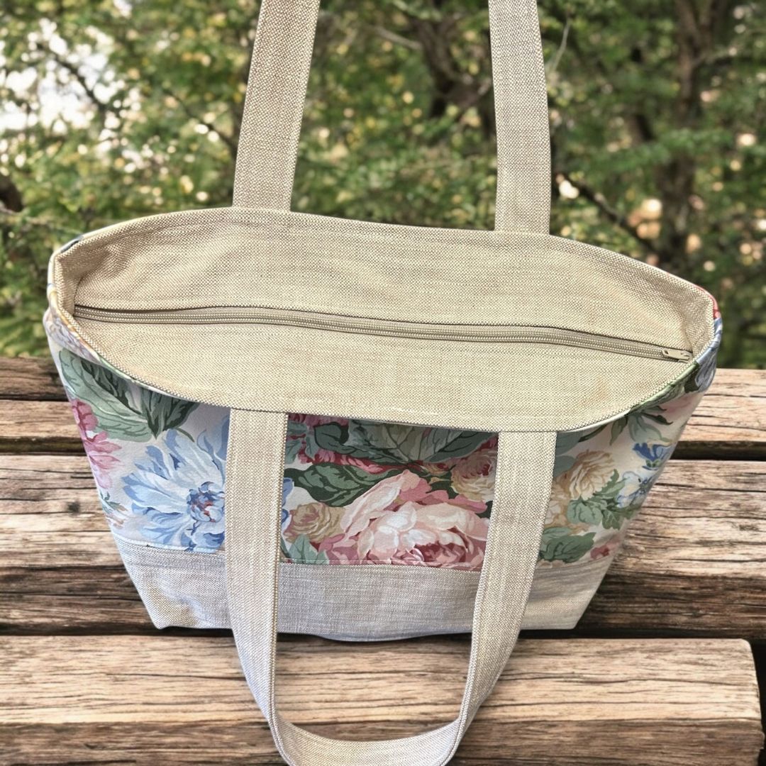 Floral tote bag with beige handles and zip closure, featuring pastel flower print and natural linen-style fabric base, photographed on a wooden bench outdoors