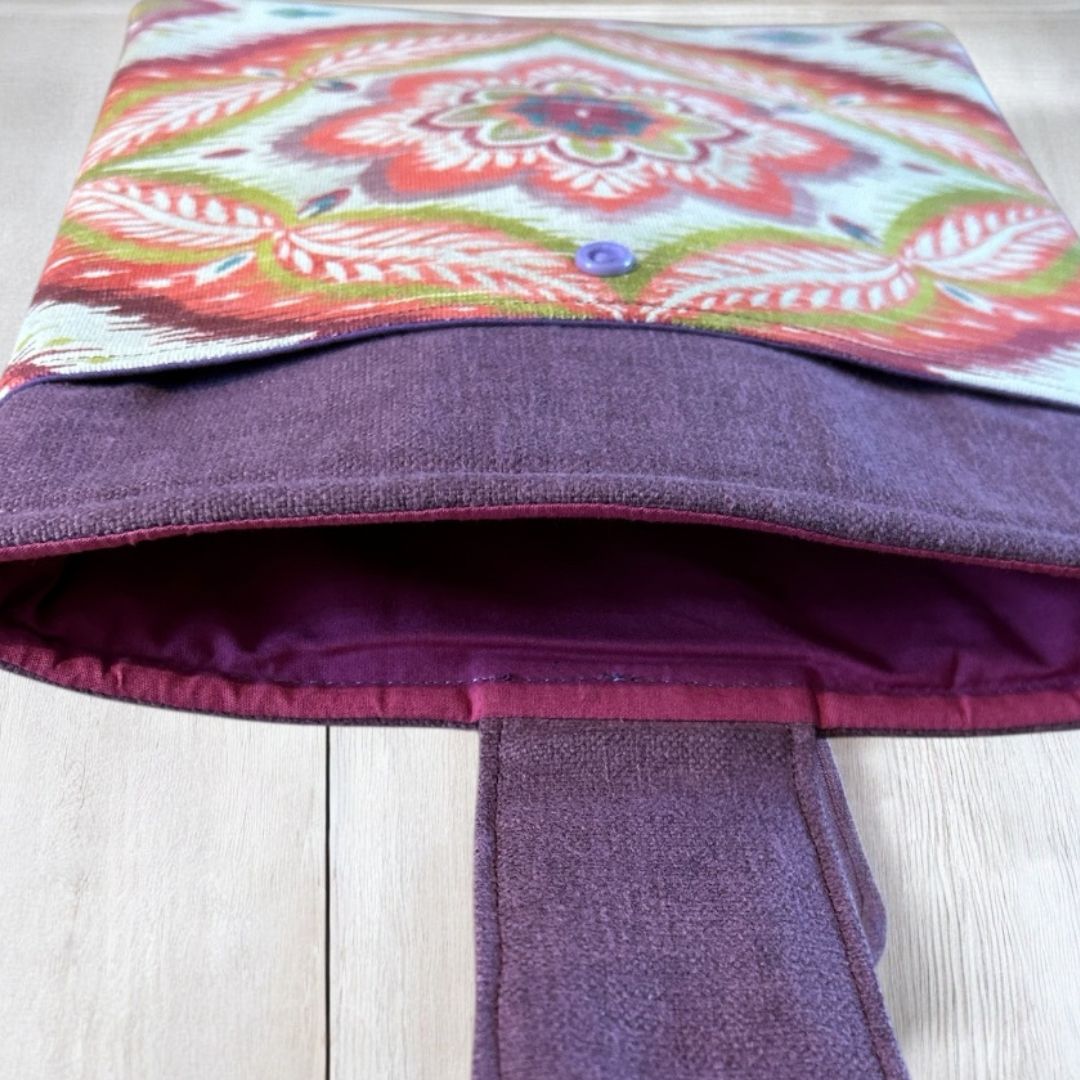 Open view of a boho tablet sleeve with red and green mandala print, plum contrast panel, burgundy lining, and snap closure on a wooden surface