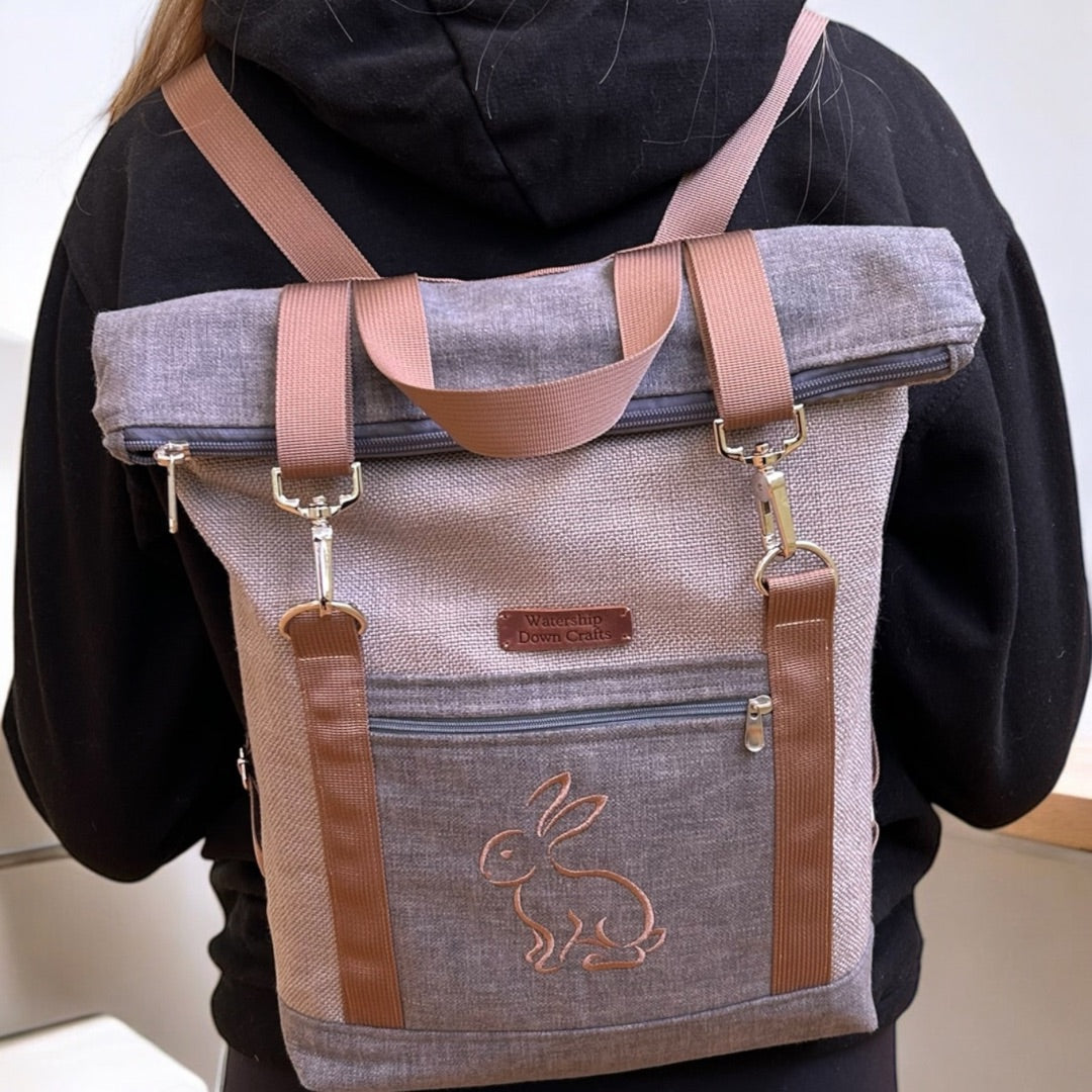 A close-up view of a roll-top backpack worn by a person in a black hoodie. The backpack is made of a neutral gray fabric with brown straps and features an embroidered outline of a rabbit on the lower front pocket. The top is rolled down and secured with silver metal clips attached to the brown straps, adding both functionality and style. A small leather patch with the "Watership Down Crafts" logo is placed near the top zipper. The combination of earthy tones and practical design gives the bag a rugged, outd