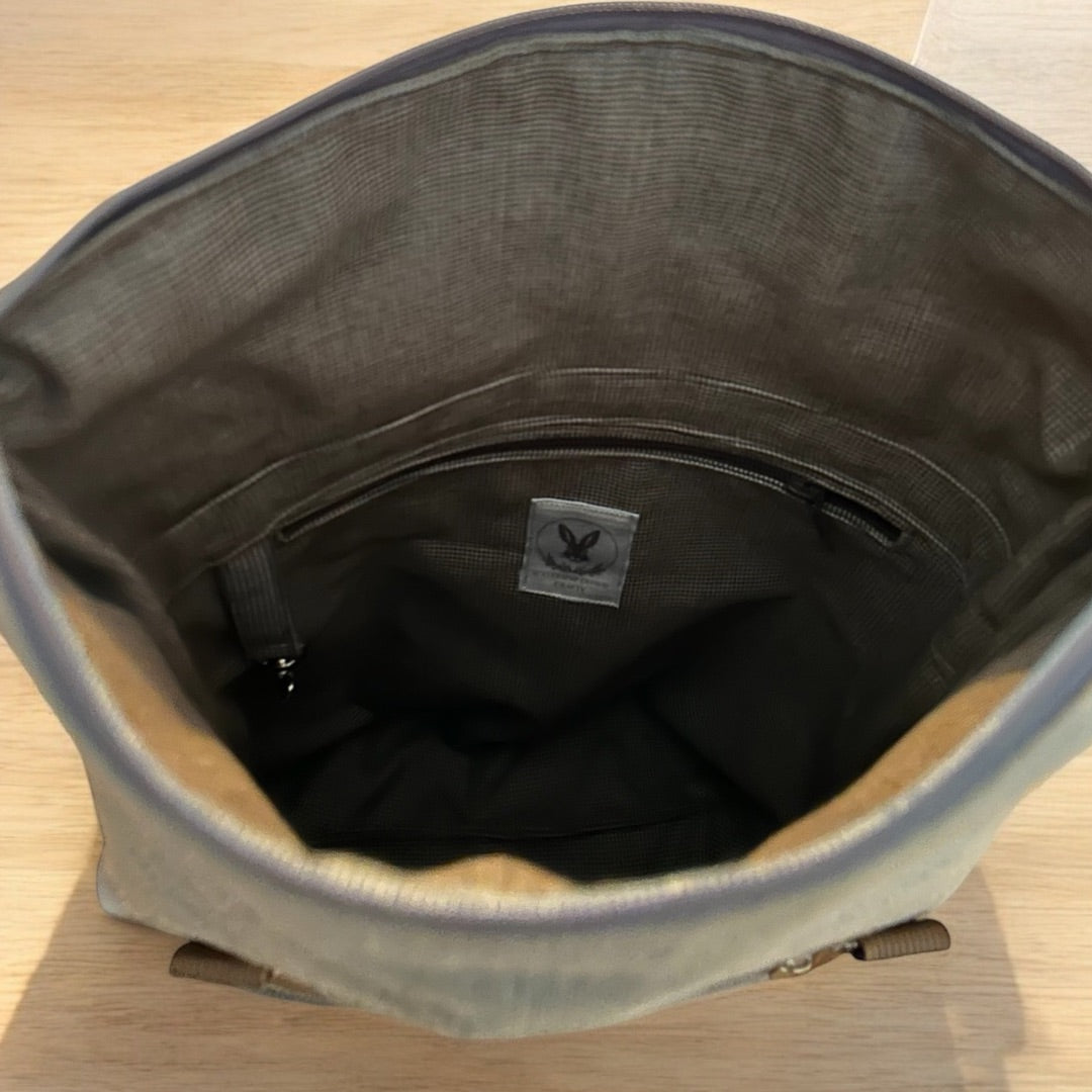 Interior view of a spacious roll-top backpack, showcasing its well-designed storage space. The lining is a muted grey with a subtle checkered texture, enhancing its sophisticated appearance. There’s a zippered pocket along the back panel for secure storage of small essentials. A label with a logo is sewn onto the lining, adding a branded touch. Near the top, a loop with a small metal clasp is visible, perfect for attaching keys or other items.