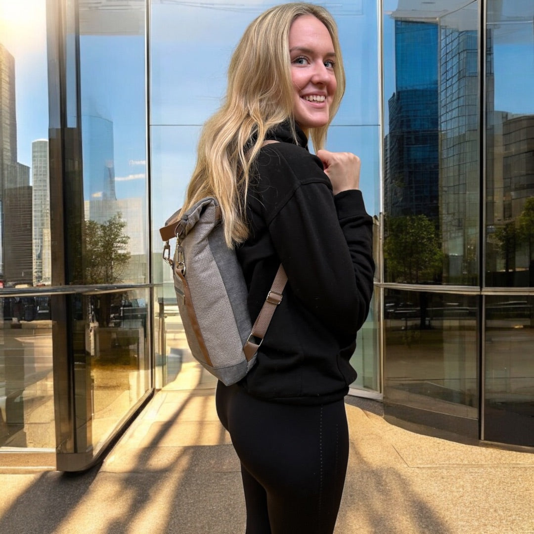 A young woman with long blonde hair wearing a black hoodie and leggings, posing outdoors with a compact roll-top backpack. The backpack has a sleek, minimalist design with grey fabric and brown straps. She stands in front of a modern glass building, smiling and looking over her shoulder, showing the backpack’s slim profile and comfortable fit. The scene captures an urban, active lifestyle, with reflections of the cityscape visible in the glass behind her.