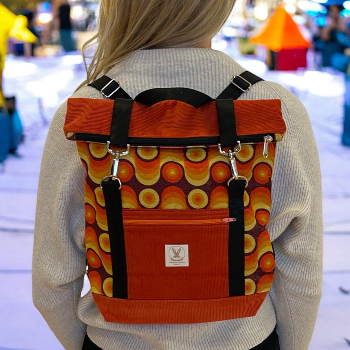 A person wearing a vibrant, retro-inspired roll-top backpack with an eye-catching orange, yellow, and brown circular pattern. The backpack features black straps and silver hardware, along with an additional orange front pocket displaying a logo tag. The wearer is dressed in a light grey sweater, and the background is softly blurred, focusing attention on the unique design and bold colors of the backpack, which evoke a vintage, 70s aesthetic.