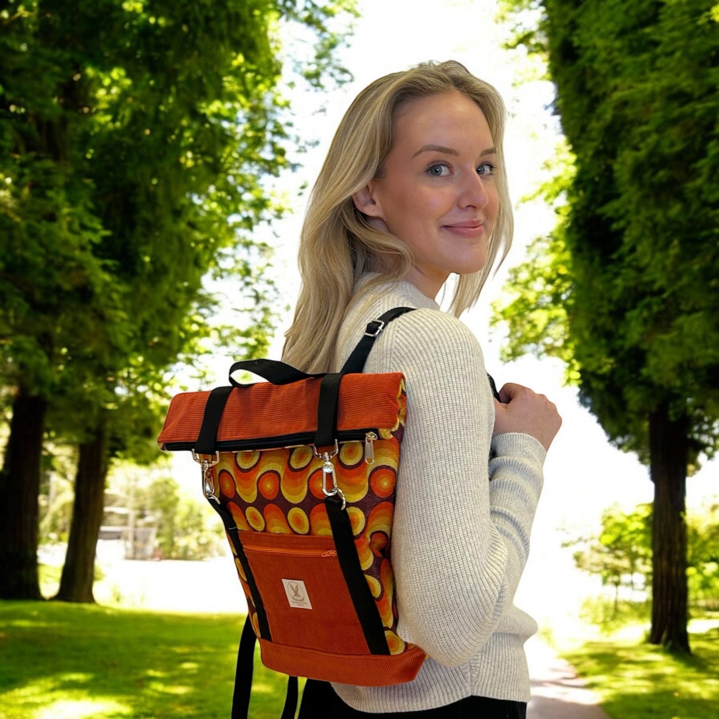 A smiling woman wearing a retro-inspired roll-top backpack in vibrant shades of orange, yellow, and brown with a bold circular pattern. The backpack has black straps and silver hardware, along with a front pocket that displays a logo tag. She is dressed in a light grey sweater and stands in a lush, sunlit park with tall trees lining the path, adding a fresh and natural backdrop that contrasts with the vintage aesthetic of the backpack.