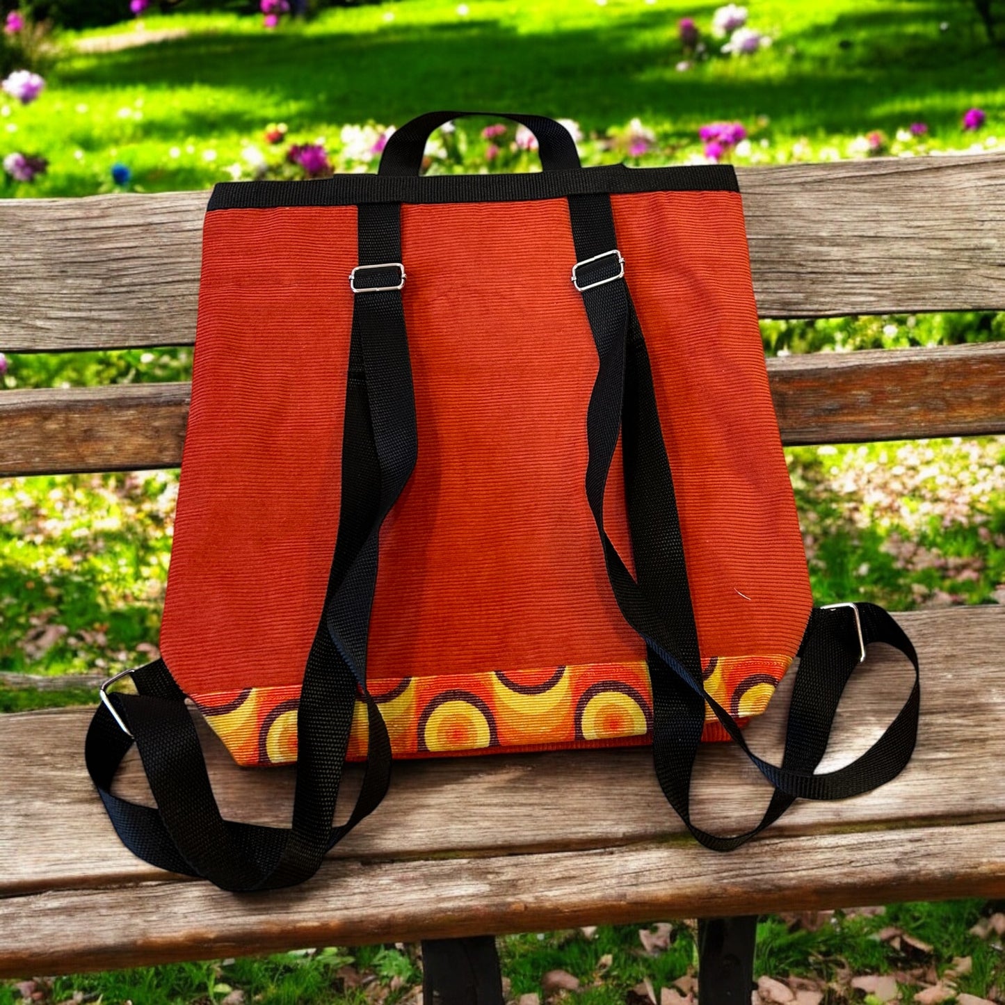A retro-inspired roll-top backpack displayed on a wooden bench in a lush garden setting with vibrant greenery and colorful flowers in the background. The backpack is crafted from orange corduroy fabric, featuring black adjustable straps and silver hardware. The bottom of the bag showcases a bold circular pattern in orange, yellow, and brown, enhancing its vintage aesthetic. The scene exudes a relaxed, natural vibe, highlighting the backpack's unique and stylish design