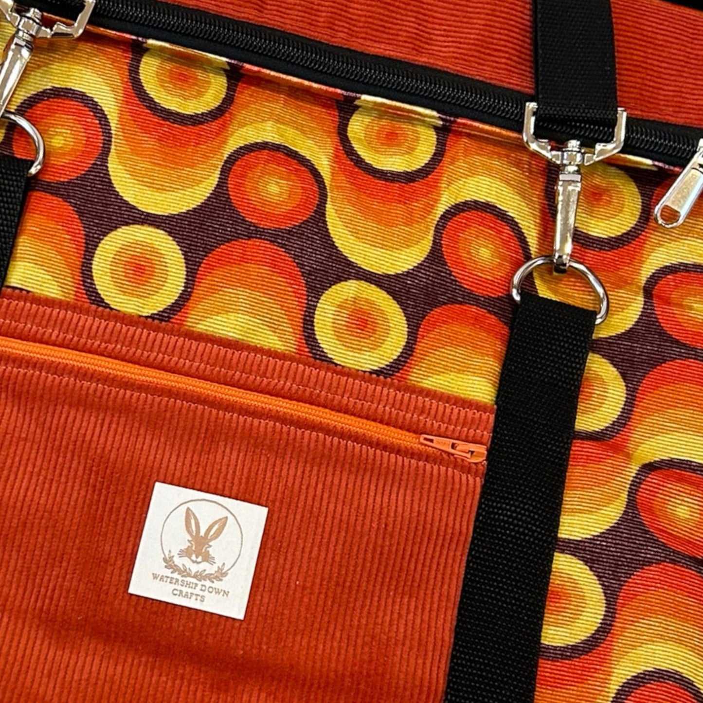 A close-up view of a retro-inspired roll-tp backpack with vibrant circular patterns in shades of orange, yellow, and brown on fabric. The bag features an orange corduroy front pocket with a matching zipper, adorned with the "Watership Down Crafts" logo patch, showcasing a rabbit emblem. Black straps with silver hardware add contrast to the design, giving it a bold, vintage look. The intricate details and vibrant colors of the pattern create a unique, eye-catching aesthetic for the backpack.