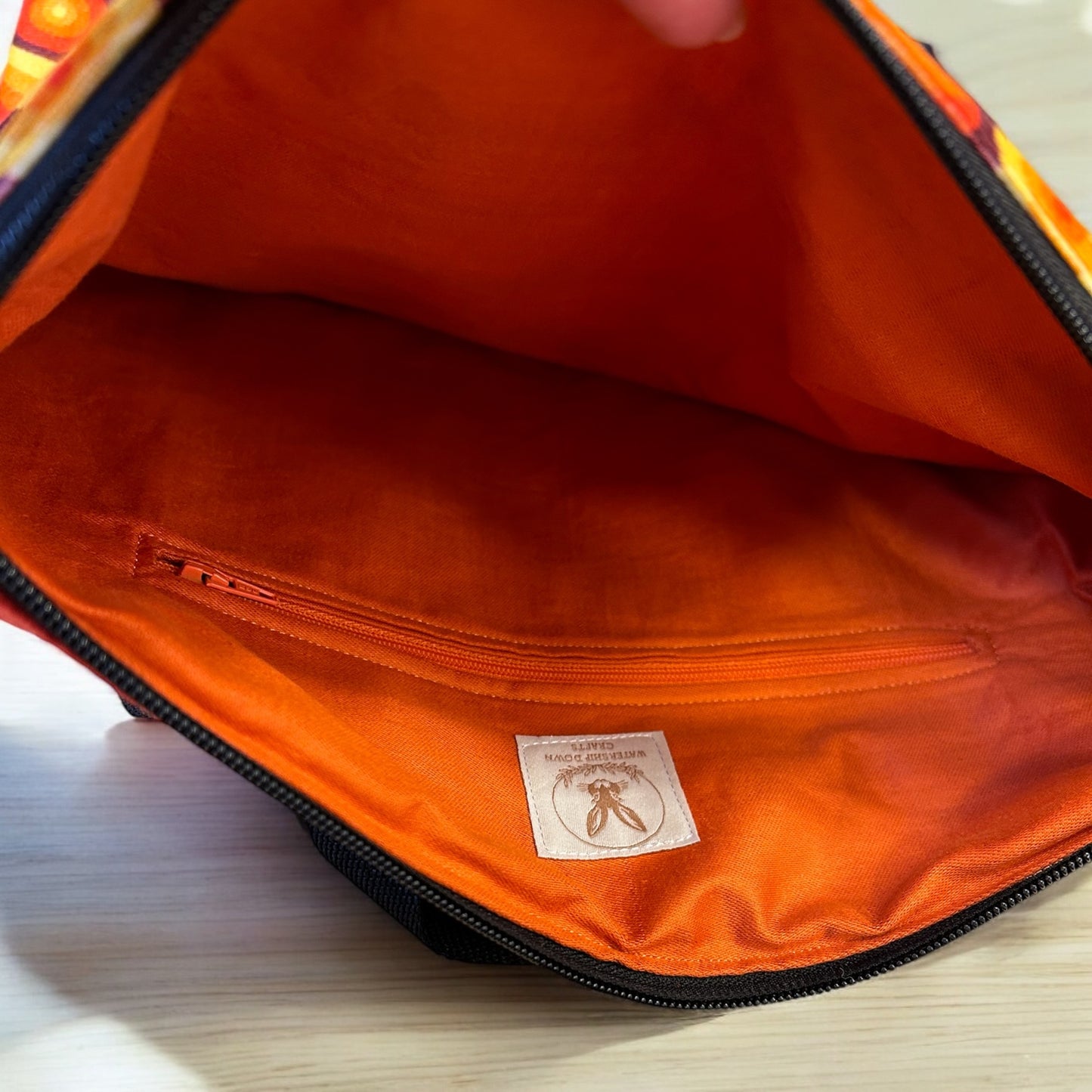 A view of the inside of a backpack with a bright orange lining, featuring an inner zipper pocket. The lining also has a fabric tag with the "Watership Down Crafts" logo, displaying a rabbit emblem. The interior is spacious and vibrant, matching the bold, retro aesthetic of the exterior design, with an orange zipper that complements the inner color scheme.