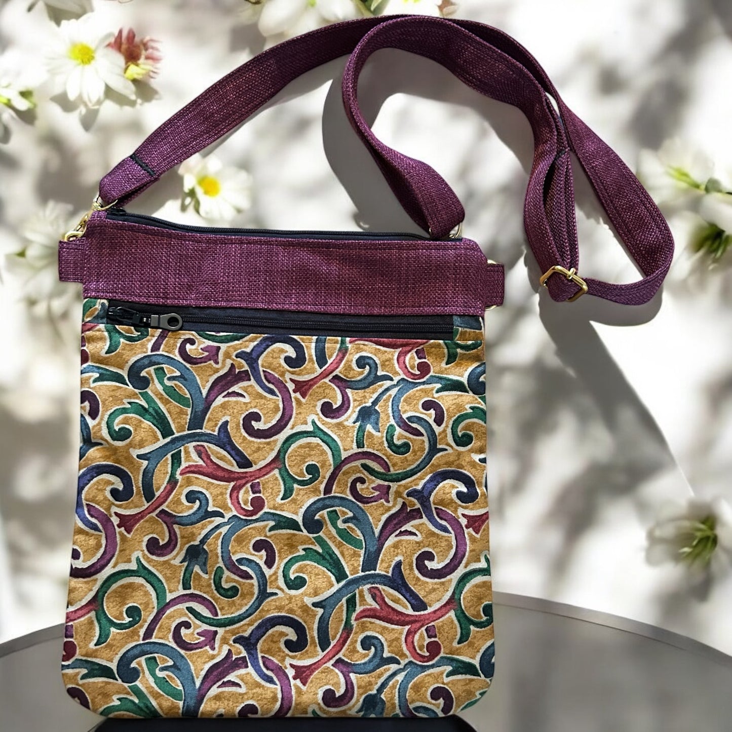 A handmade crossbody bag with a vibrant multicolored swirl pattern on the main body, accented with a deep purple fabric at the top and a matching adjustable strap. The bag features a zip closure, combining a practical and stylish design, set against a soft floral background with natural lightin