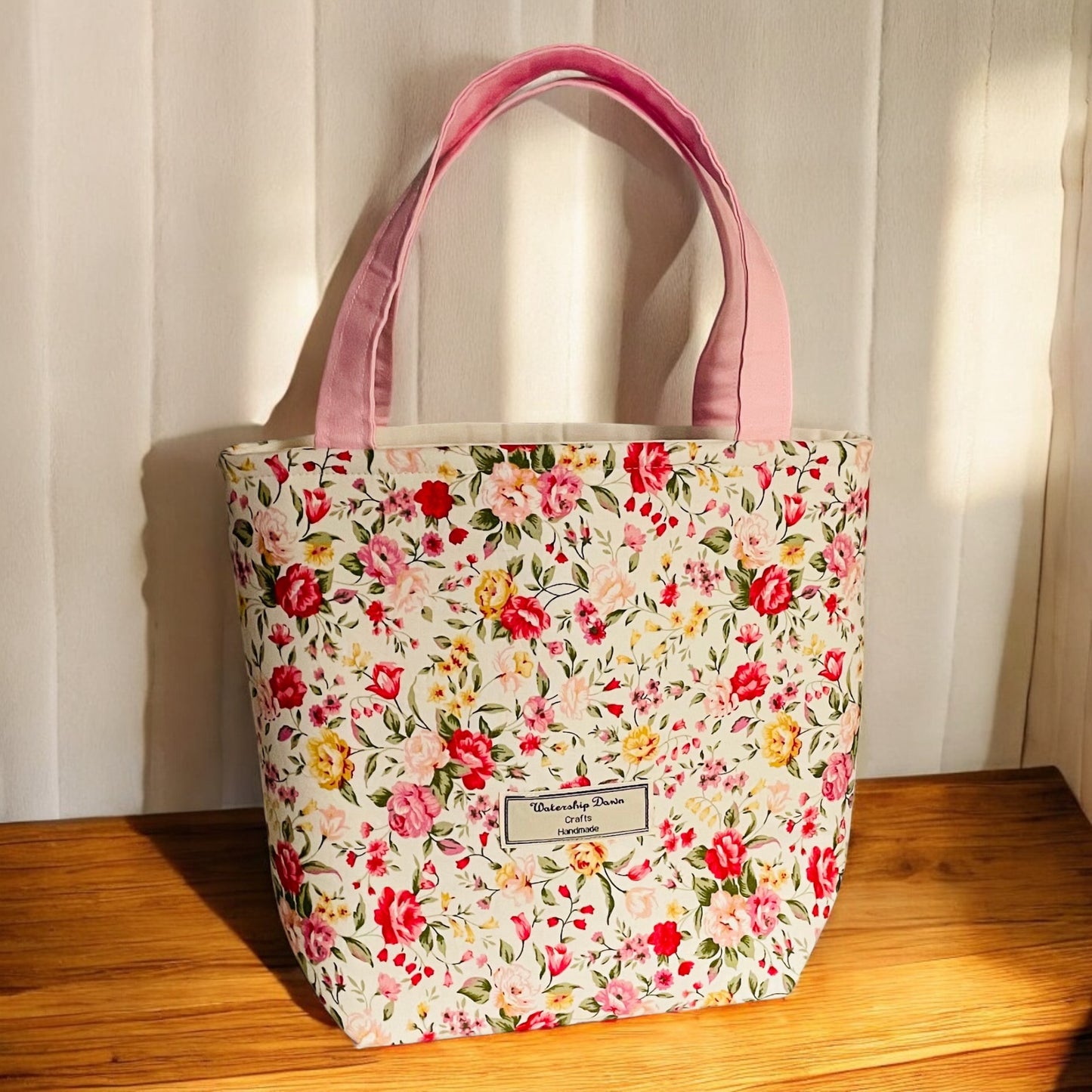 A beautifully handmade mini tote bag with a soft floral print featuring pink, red, and yellow flowers on a cream background. The bag is accented with light pink fabric handles, adding a delicate touch to its charming design. A "Watership Down Crafts Handmade" label is stitched on the front, emphasizing the bag's handcrafted quality. It is displayed in soft sunlight against a light wooden surface, creating a fresh and inviting look.