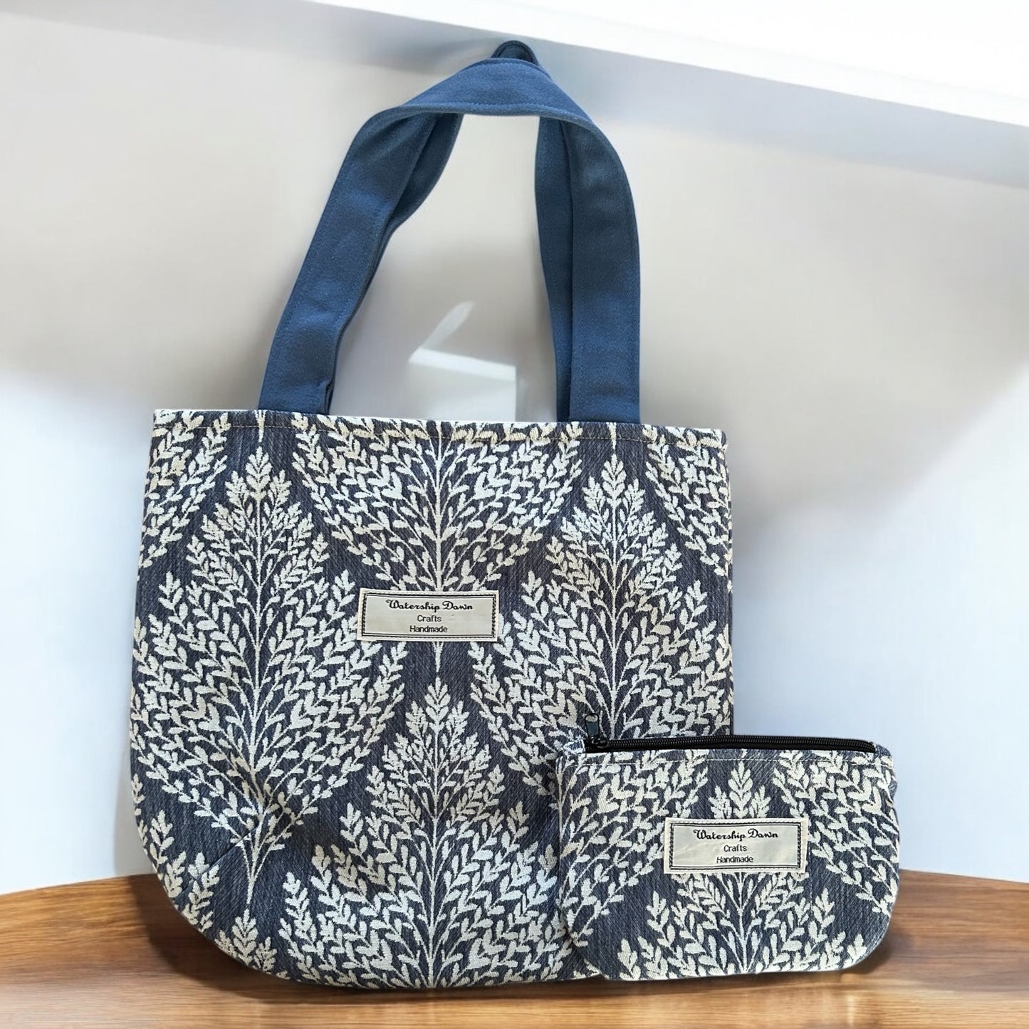 A beautifully coordinated set of handmade accessories, featuring a tote bag and matching pouch. The tote bag has a bold, nature-inspired leaf pattern in white against a dark blue background, complemented by sturdy blue handles. The matching pouch features the same design, providing a convenient and stylish way to organize smaller items. Both items prominently display the "Watership Down Crafts Handmade" label, showcasing their handcrafted quality. The set is displayed against a clean, minimalist background,