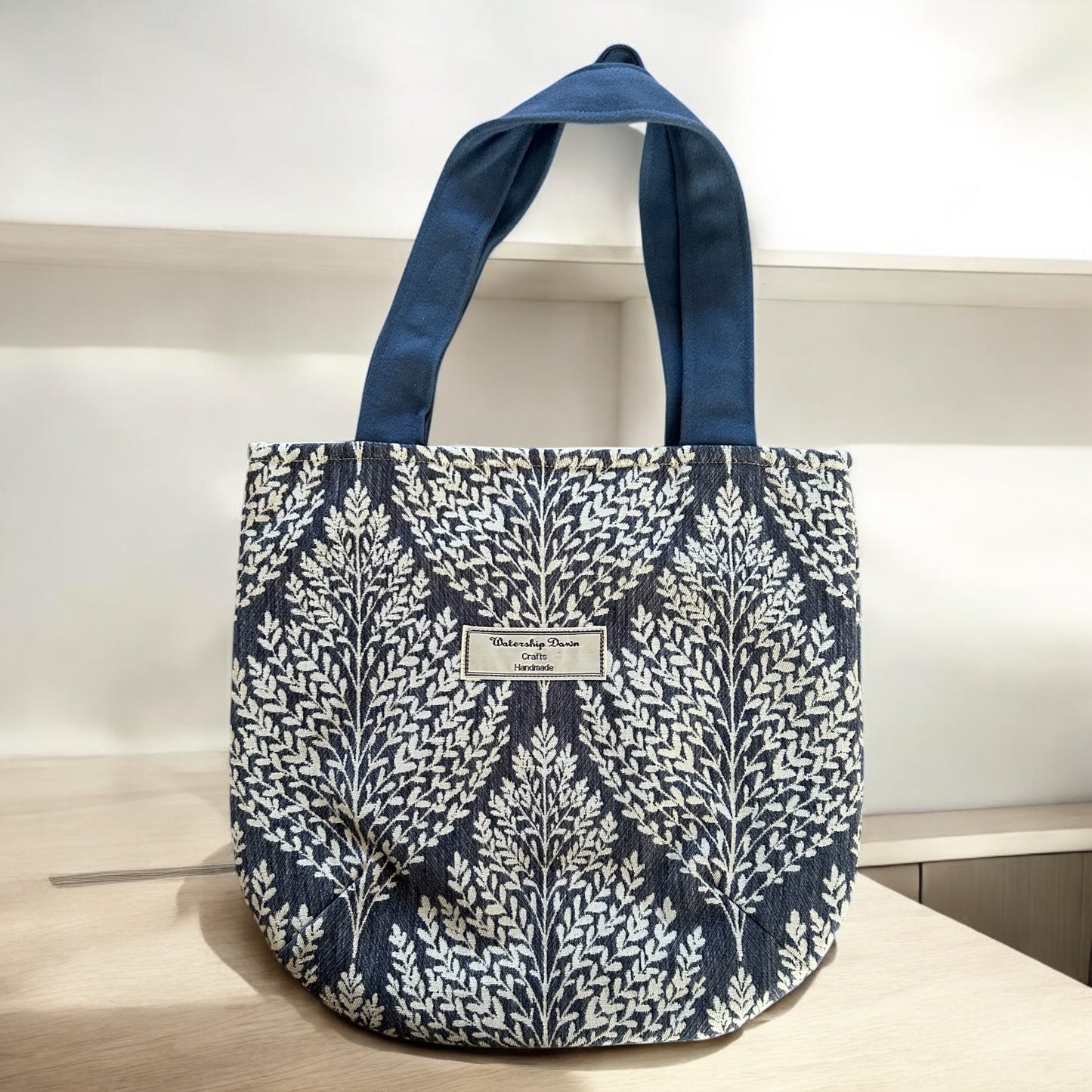 A handmade tote bag featuring an elegant botanical design with intricate white leaf patterns against a dark blue background. The bag is complemented by blue fabric handles that enhance the overall sophisticated look. The "Watership Down Crafts Handmade" label is sewn onto the front, adding a touch of authenticity. It is displayed against a clean, minimalist background, highlighting its stylish and natural aesthetic.