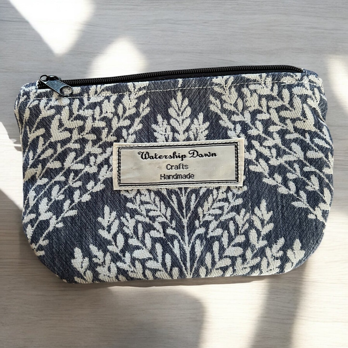 A handmade zip pouch featuring an elegant botanical print with intricate white leaf patterns set against a dark blue background. The pouch is accented with a "Watership Down Crafts Handmade" label on the front, emphasizing its handcrafted nature. The design is both stylish and practical, perfect for organizing small items while maintaining a modern, nature-inspired aesthetic. The pouch is photographed in soft natural light, highlighting the detailed fabric pattern.