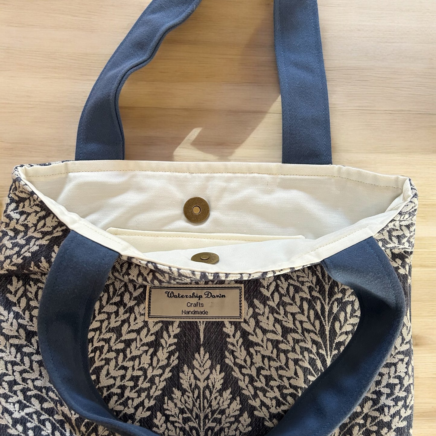 A close-up of the interior of a handmade tote bag, showcasing a clean, neutral lining with a magnetic snap closure for added security. The bag's exterior features a botanical leaf print in white against a dark blue background. The bag's "Watership Down Crafts Handmade" label is visible inside, and the sturdy blue fabric handles add a contrasting touch. The design is functional and stylish, combining practical features with an elegant botanical aesthetic.