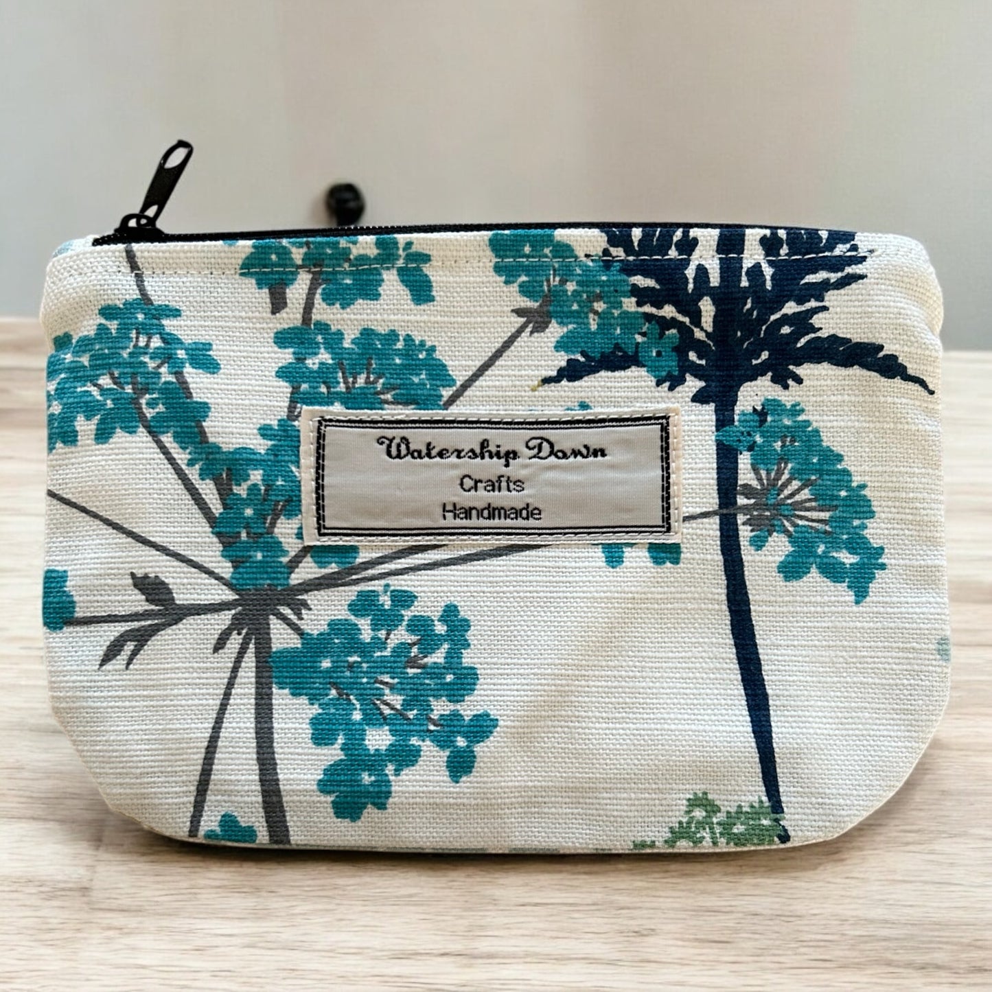 A handmade zip pouch featuring a botanical print with teal and navy flowers on a neutral cream background. The pouch is accented with a "Watership Down Crafts Handmade" label, highlighting its handcrafted quality. The design is clean and functional, perfect for organizing small items while maintaining a stylish and modern aesthetic. The pouch is photographed against a light wooden surface, showcasing its fresh and nature-inspired design.