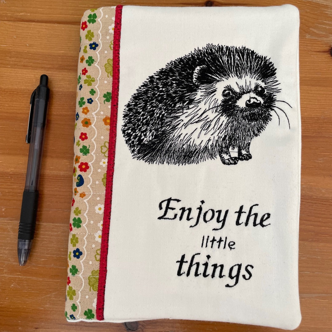 Handmade notebook cover featuring an embroidered hedgehog design and the phrase 'Enjoy the little things' in elegant script. The cover includes a decorative floral fabric strip on the left, bordered by a red accent, showcasing a rustic and cozy aesthetic.