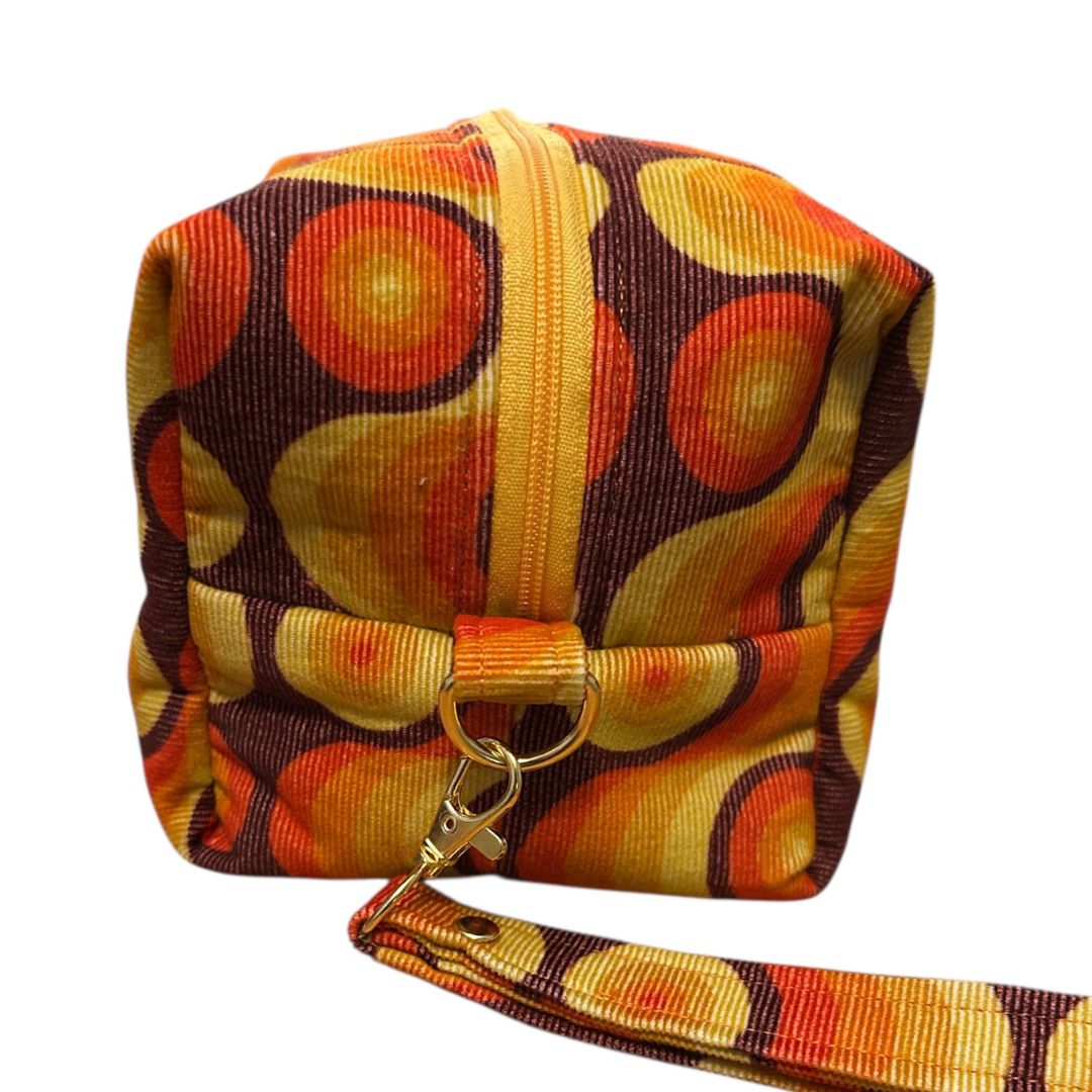Side view of handmade retro makeup bag with yellow zip, detachable wrist strap, and gold swivel clasp. Fabric features bold orange, yellow, and brown swirl pattern.