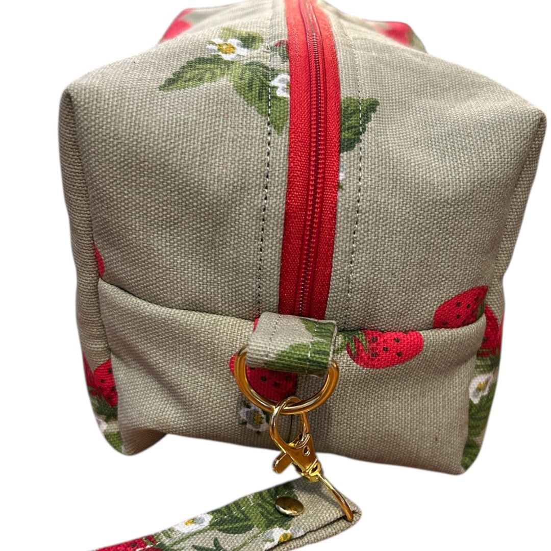 Side view of strawberry print makeup bag showing detachable wrist strap with gold hardware, red zip, and boxed corner detail.