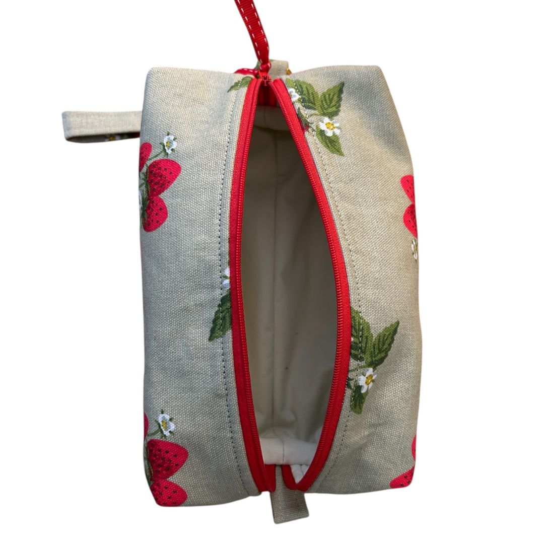 Top-down view of open handmade makeup bag showing cream water-resistant lining, red zip, and Sophie Allport strawberry fabric exterior.
