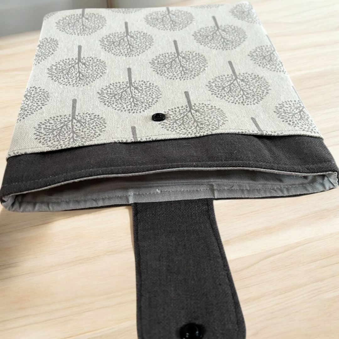 inside view of a beige and dark grey tree print tablet case with snap closure, showing soft padded lining and flap open on a light wooden surface