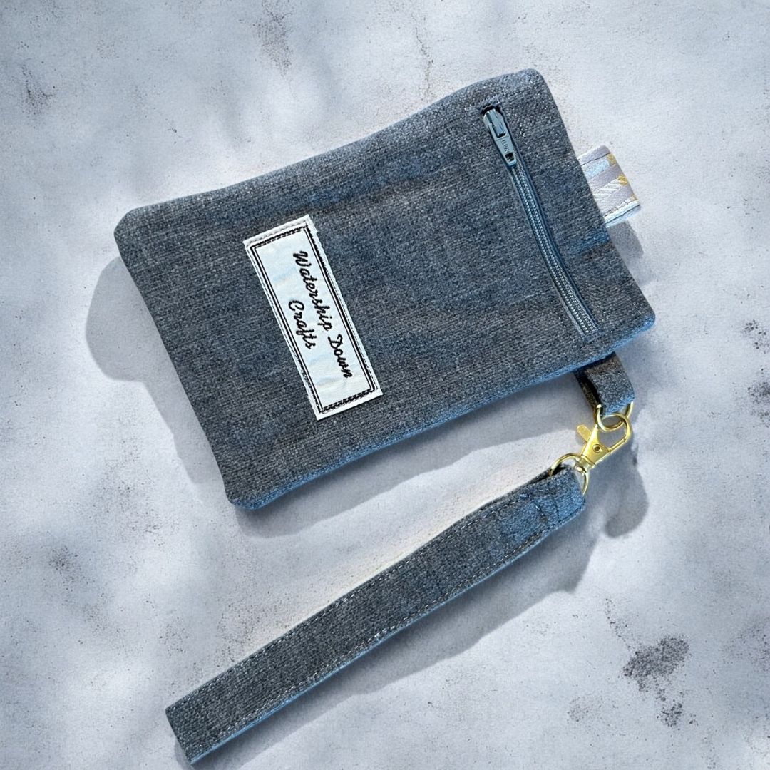 Back view of a handmade phone case in charcoal grey with zip-up pocket, detachable wristlet, and Watership Down Crafts label