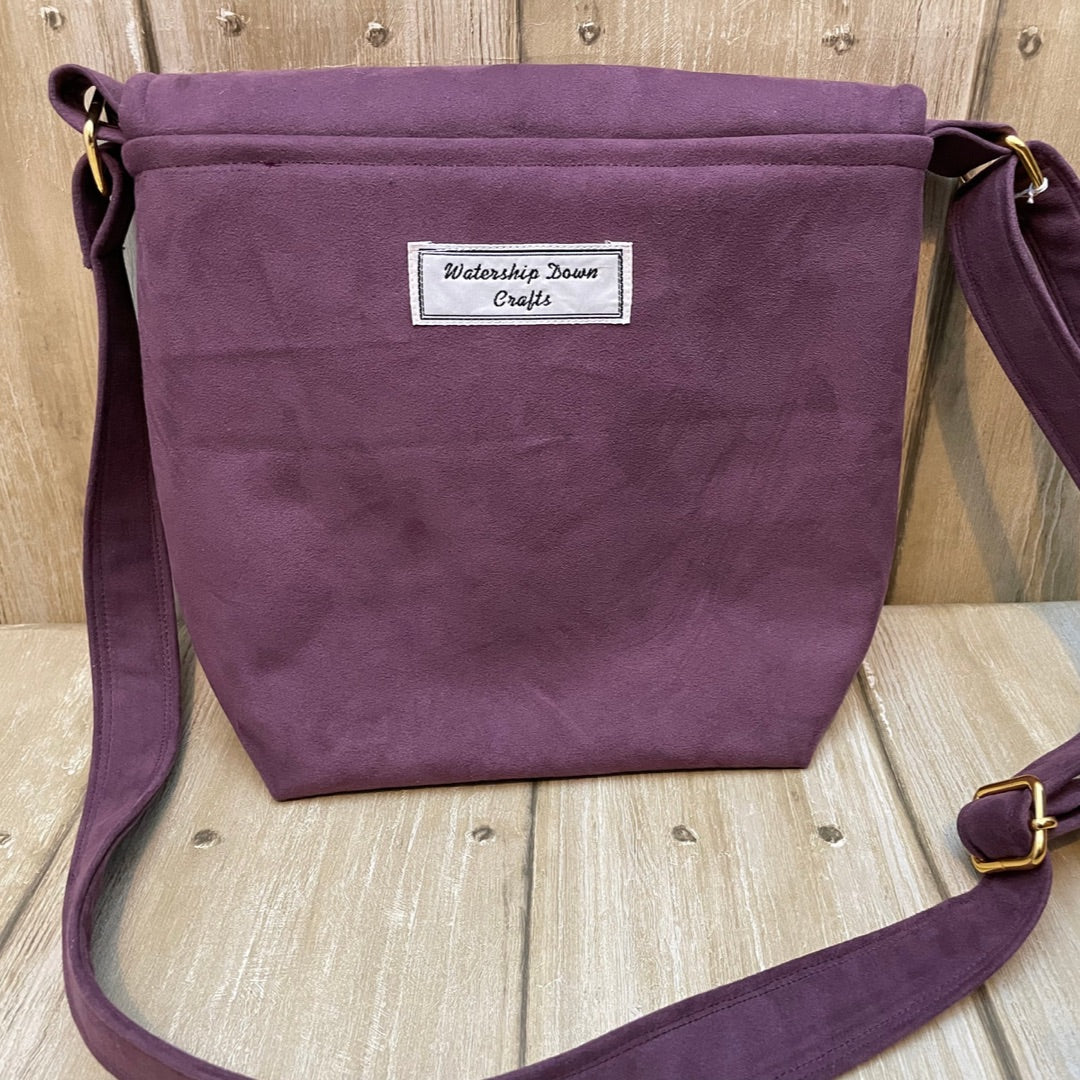 The back view of a maroon shoulder bag displayed against a rustic wooden background. The bag features a simple design with a white fabric label that reads "Watership Down Crafts" stitched near the top center. It has a matching maroon strap with adjustable metal hardware on the side, allowing for easy carrying. The fabric has a soft, suede-like texture, giving the bag a refined, handmade look.