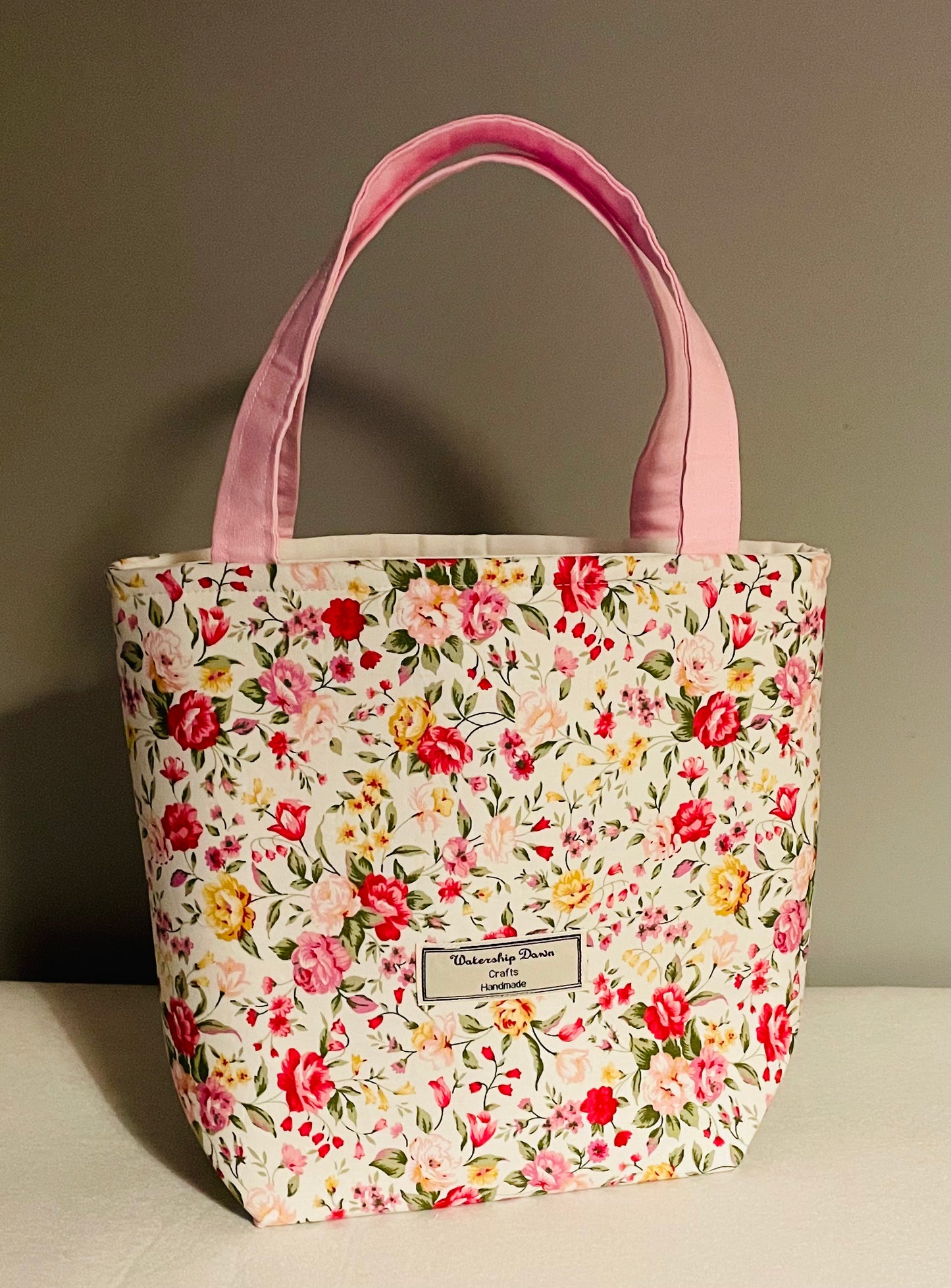 A charming handmade mini tote bag featuring a vibrant floral design with pink, red, and yellow flowers on a soft cream background. The bag is completed with light pink handles that complement the floral pattern beautifully. A "Watership Down Crafts Handmade" label is sewn onto the front, emphasizing its crafted quality. The bag is displayed against a neutral backdrop, highlighting its bright and feminine design.