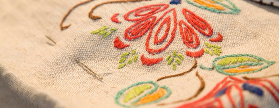 Close-up of a hand-embroidered floral design in vibrant red, green, and orange threads on cream fabric, with a needle still in place.