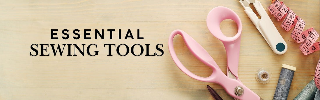 Flat lay of essential sewing tools including pink fabric scissors, seam ripper, measuring tape and thread spools arranged on a wooden surface.