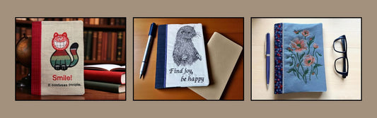 Three embroidered A5 notebook covers displayed side by side: one with a smiling cat and the words “Smile! It confuses people,” one with an otter and the text “Find joy, be happy,” and one with embroidered flowers on a blue cover.