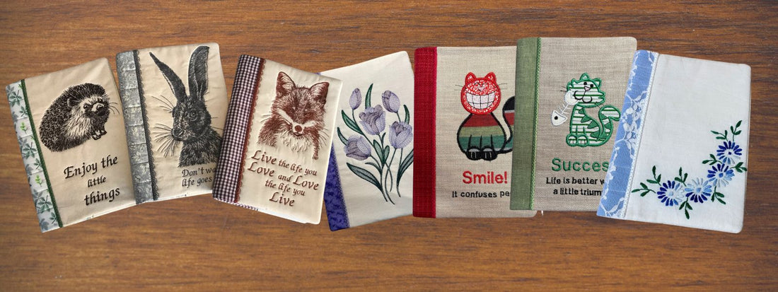 A row of seven embroidered notebook covers laid out on a wooden surface, featuring woodland animals, flowers, inspirational quotes, and colourful fabric spines.”