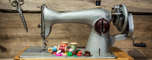 A vintage grey sewing machine sits on a wooden table, with colourful spools of thread, buttons, and sewing accessories placed in front. Scissors hang on a wooden background, adding to the rustic aesthetic of the scene.