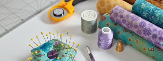 A sewing workspace featuring colorful fabric rolls, a rotary cutter, thread spools, a pincushion with yellow-headed pins, and a thimble on a cutting mat—essential tools for sewing projects.