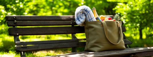 An olive-green tote bag sits on a wooden park bench, filled with fresh produce, a rolled-up picnic blanket, and other essentials. The background features lush green trees and sunlight filtering through the leaves.