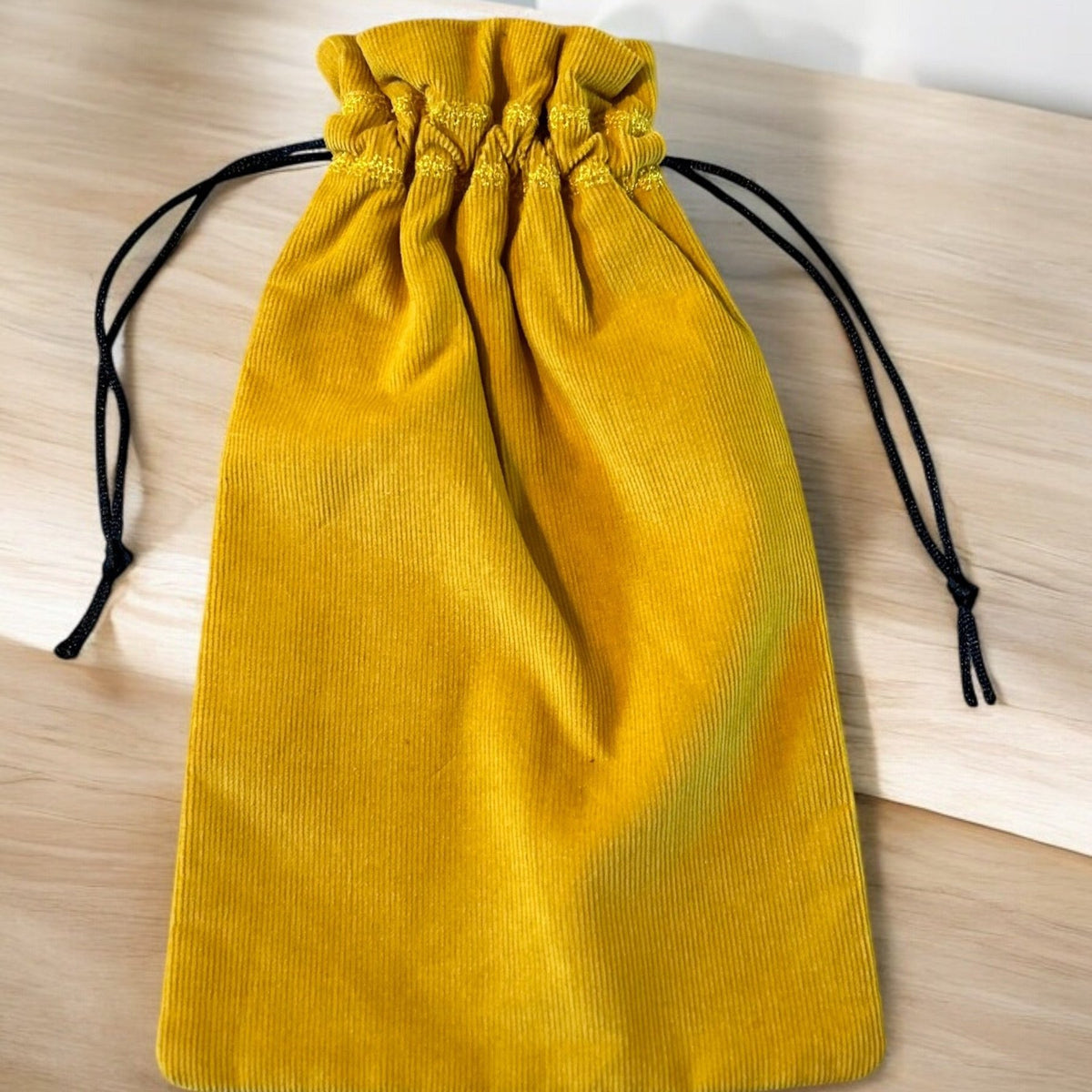 A handmade drawstring bag crafted from mustard yellow corduroy fabric, showcasing a soft textured appearance. The bag is cinched with black drawstring cords for a contrasting touch, combining functionality with bold color. It is displayed on a light wooden surface, highlighting the richness of the fabric.