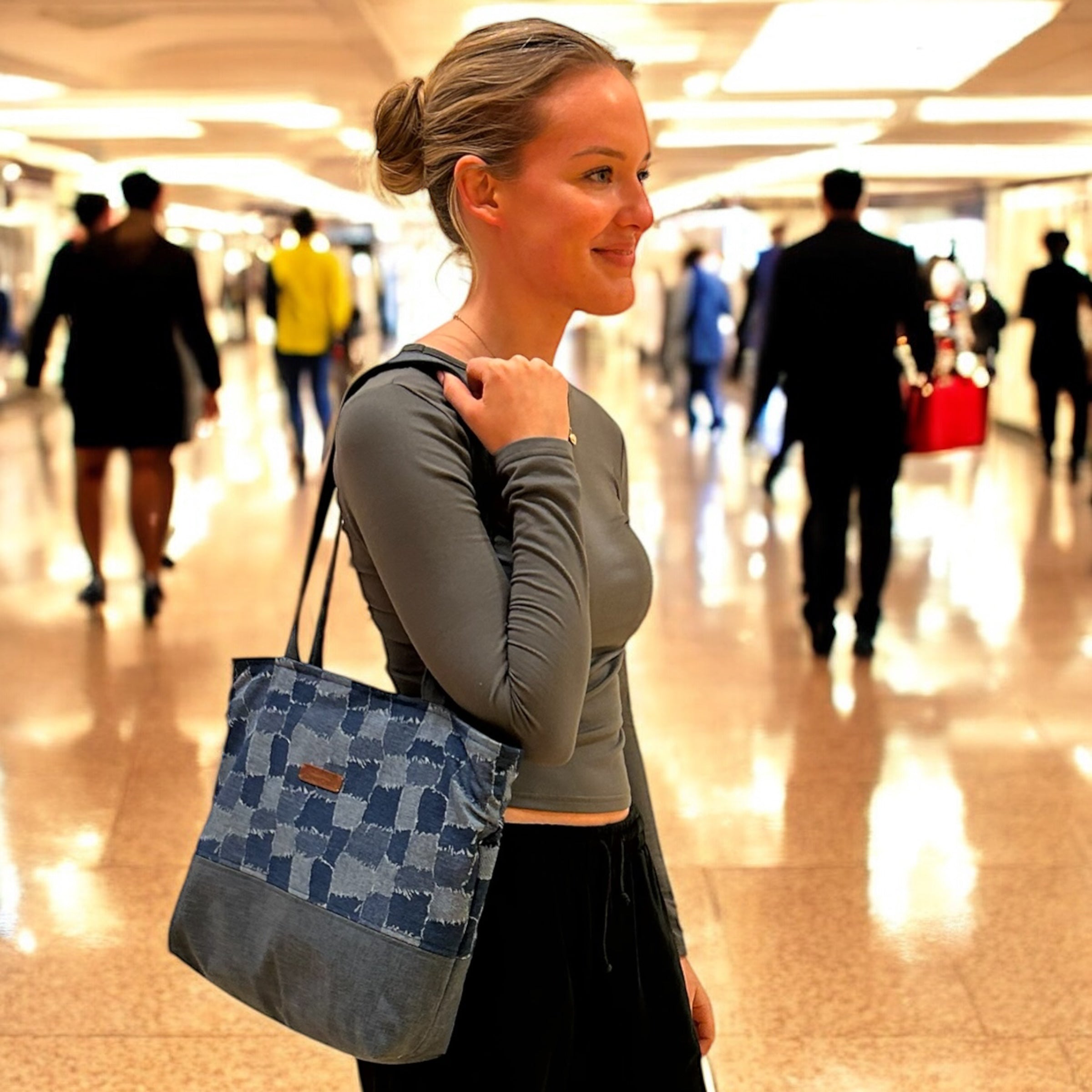 Smiling woman standing in a busy indoor walkway, holding a denim patchwork tote bag over her shoulder. The bag features a checkerboard design with contrasting fabric panels, blending style and practicality for an on-the-go lifestyle.