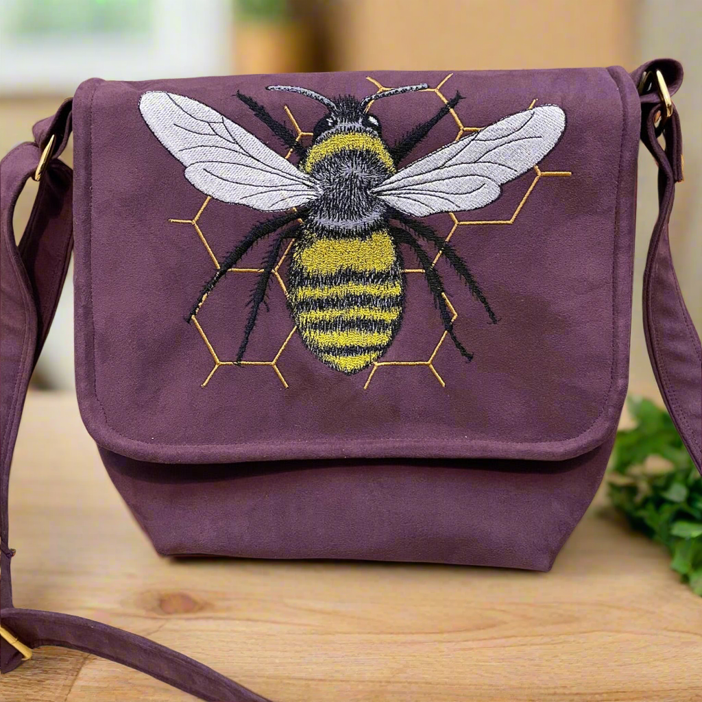 A maroon shoulder bag with an embroidered design of a bee on the front flap. The bee features detailed stitching in yellow, black, and white, with intricate wings and a fuzzy body, set against a background of honeycomb outlines in gold thread. The bag has a rustic, handcrafted appearance and includes a matching maroon strap with metal hardware. The bag is displayed on a wooden surface, adding to its earthy, artisanal charm.