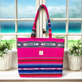 Vibrant handmade pink and blue boho tote bag with intricate woven patterns, displayed on a windowsill overlooking a serene beach view. Featuring long handles and a spacious design, this bag blends practicality with bold, colorful style.