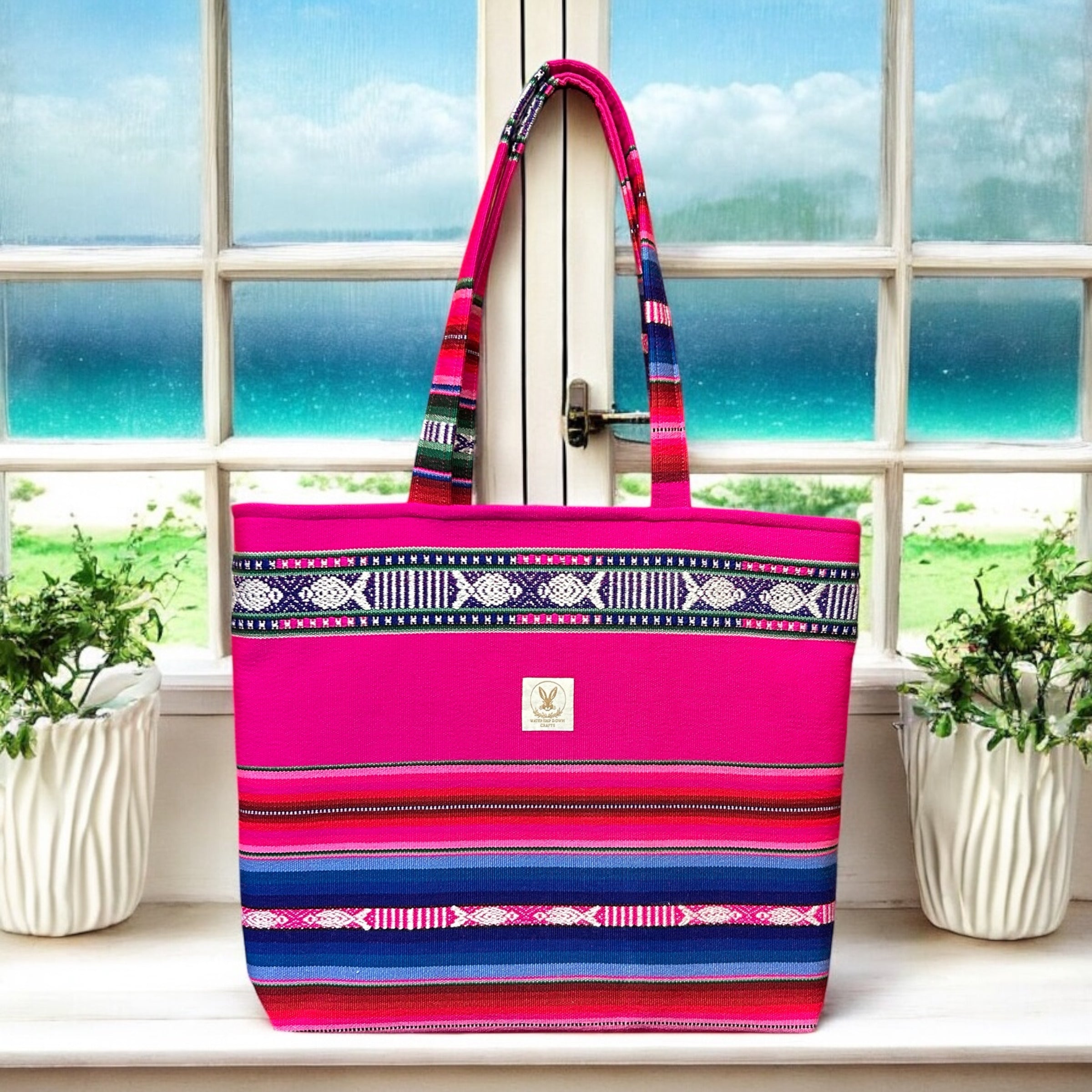 Vibrant handmade pink and blue boho tote bag with intricate woven patterns, displayed on a windowsill overlooking a serene beach view. Featuring long handles and a spacious design, this bag blends practicality with bold, colorful style.